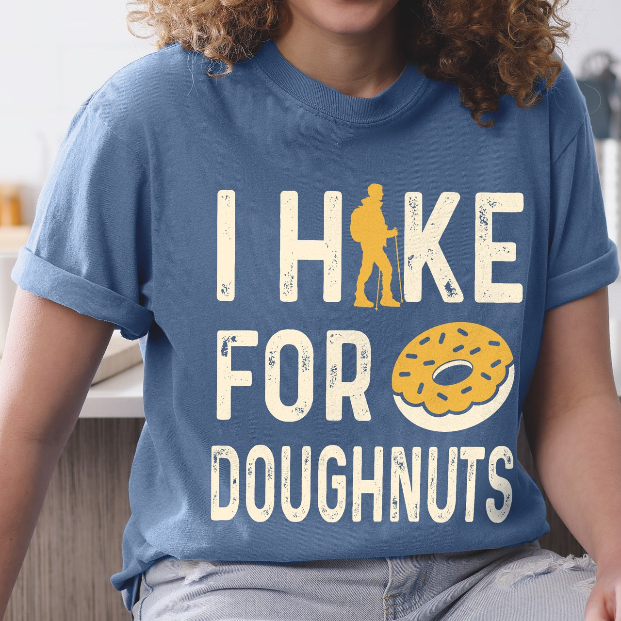 Woman wearing Blue Jean (blue) Doughnut donut lover statement tee I Hike for Doughnuts with vintage distressed cream typography character silhouette with trekking pole and illustrated glazed doughnut with sprinkles on Comfort Colors shirt for sweet treat enthusiasts, hikers and foodies.