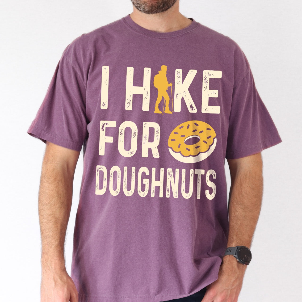 Man wearing Berry (purple) Doughnut donut lover statement tee I Hike for Doughnuts with vintage distressed cream typography character silhouette with trekking pole and illustrated glazed doughnut with sprinkles on Comfort Colors shirt for sweet treat enthusiasts, hikers and foodies.