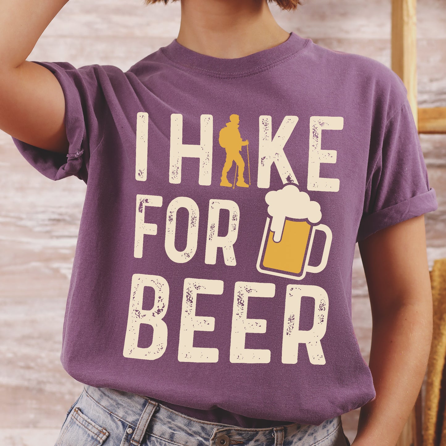 Berry (Dark Violet) graphic t-shirt with 'I Hike for Beer' text in cream lettering featuring orange hiker silhouette and beer mug illustration with foam, styled with light wash jeans and brown hiking boots