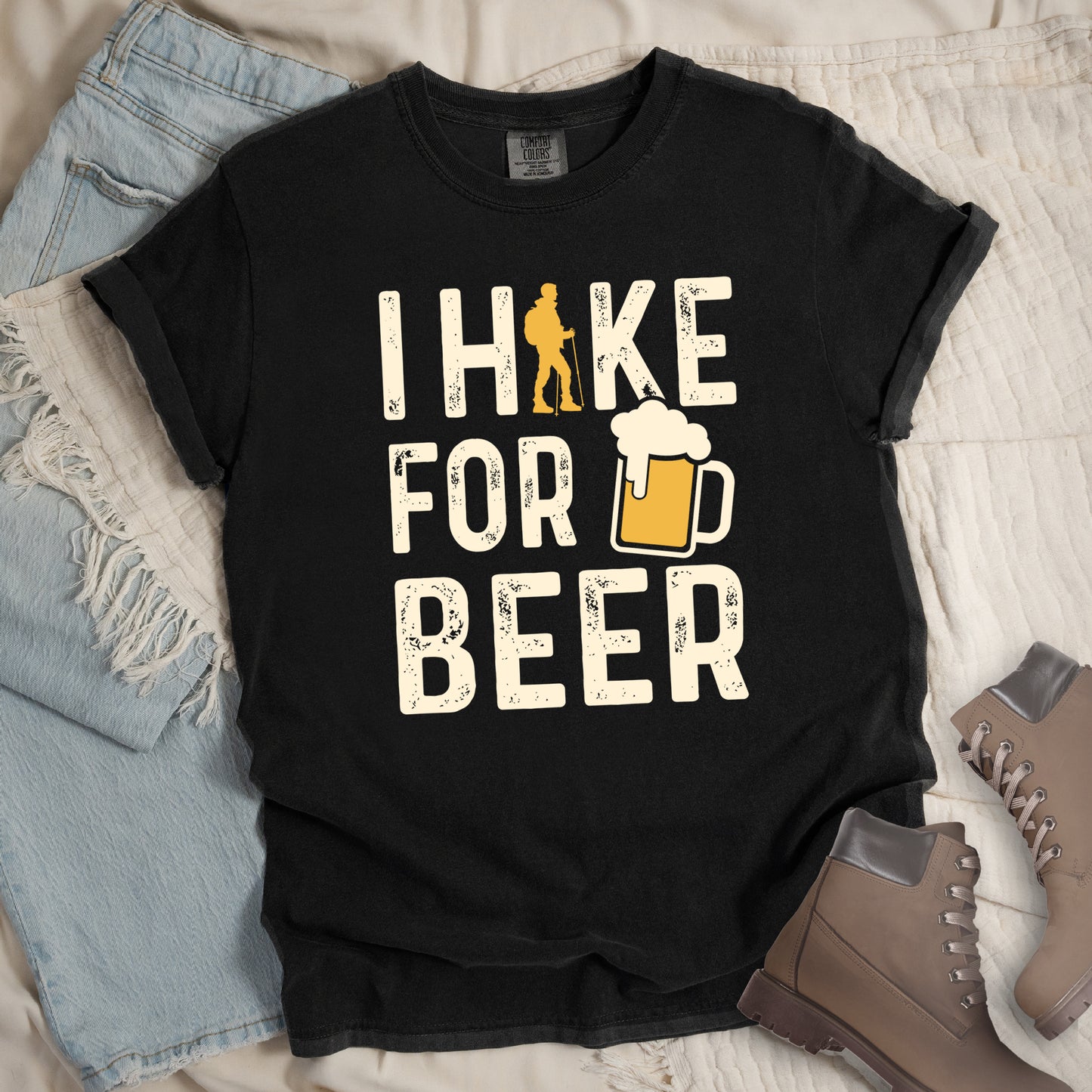 Black graphic t-shirt with 'I Hike for Beer' text in cream lettering featuring orange hiker silhouette and beer mug illustration with foam, styled with light wash jeans and brown hiking boots.