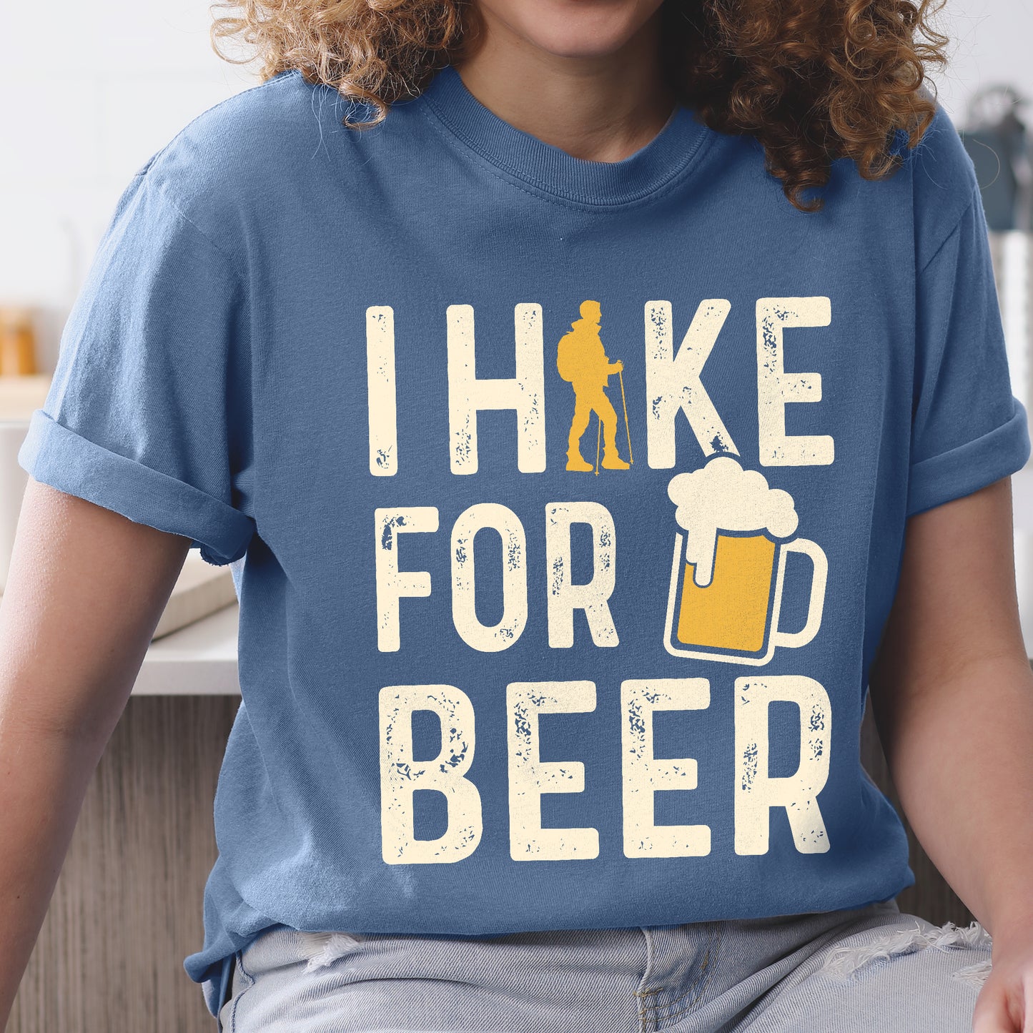 Blue Jean (Denim Blue) graphic t-shirt with 'I Hike for Beer' text in cream lettering featuring orange hiker silhouette and beer mug illustration with foam, styled with light wash jeans and brown hiking boots