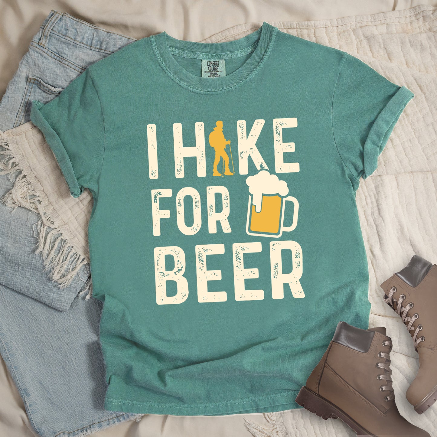 Light Green graphic t-shirt with 'I Hike for Beer' text in cream lettering featuring orange hiker silhouette and beer mug illustration with foam, styled with light wash jeans and brown hiking boots.