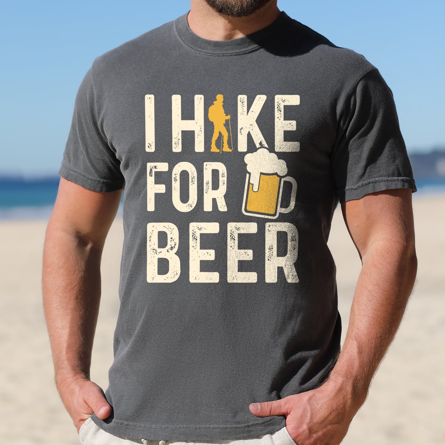 Pepper (Dark Gray) graphic t-shirt with 'I Hike for Beer' text in cream lettering featuring orange hiker silhouette and beer mug illustration with foam, styled with light wash jeans and brown hiking boots
