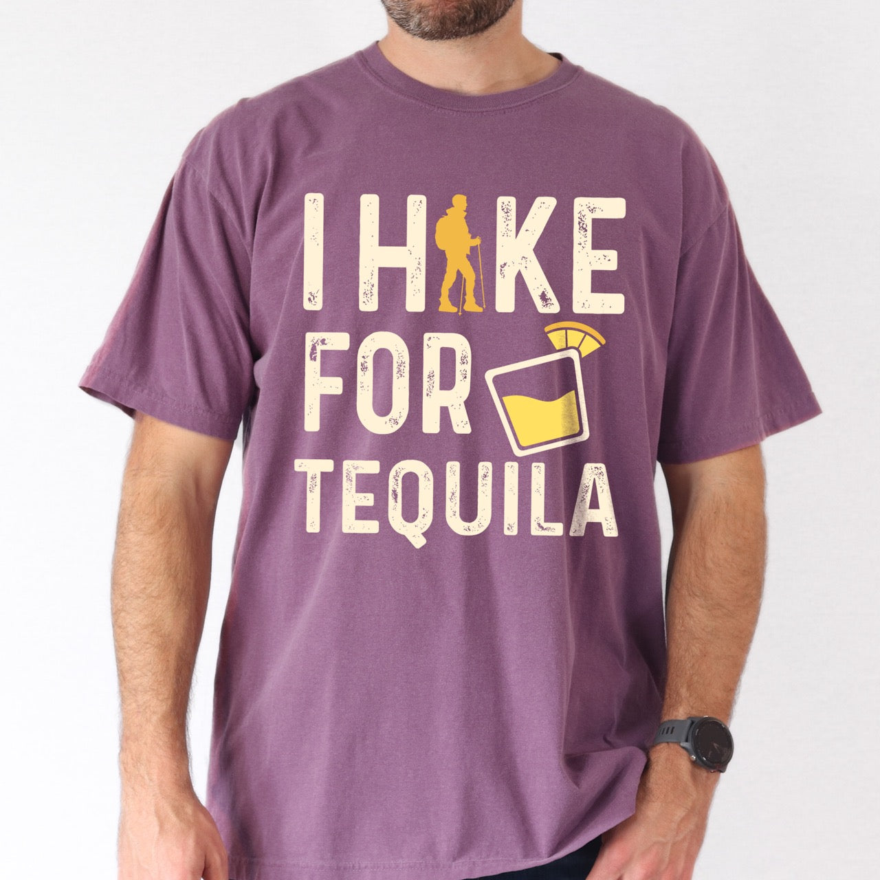 Man wearing Berry (purple) Tequila margarita lover statement tee I Hike for Tequila with vintage distressed cream typography character silhouette with trekking pole and illustrated tilted margarita glass with lime garnish on Comfort Colors shirt for drink enthusiasts for hikers and foodies.