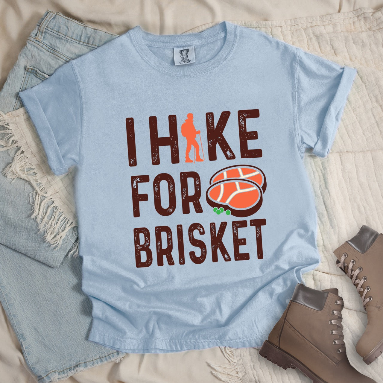 Chambray (light blue) BBQ brisket lover statement tee I Hike for Brisket with distressed brown typography character silhouette with trekking pole and illustrated sliced smoked brisket with herb garnish on vintage Comfort Colors shirt for food motivated adventurers for hikers and foodies - laid flat with jeans and boots.