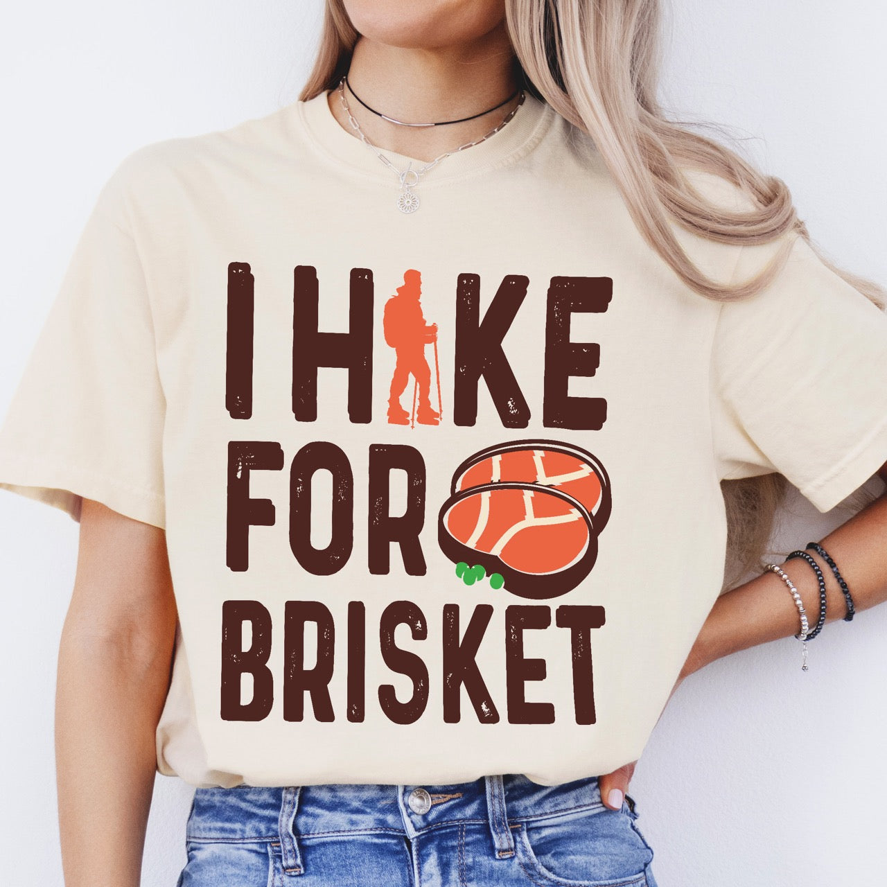 Woman wearing Ivory BBQ brisket lover statement tee I Hike for Brisket with distressed brown typography character silhouette with trekking pole and illustrated sliced smoked brisket with herb garnish on vintage Comfort Colors shirt for food motivated adventurers for hikers and foodies.