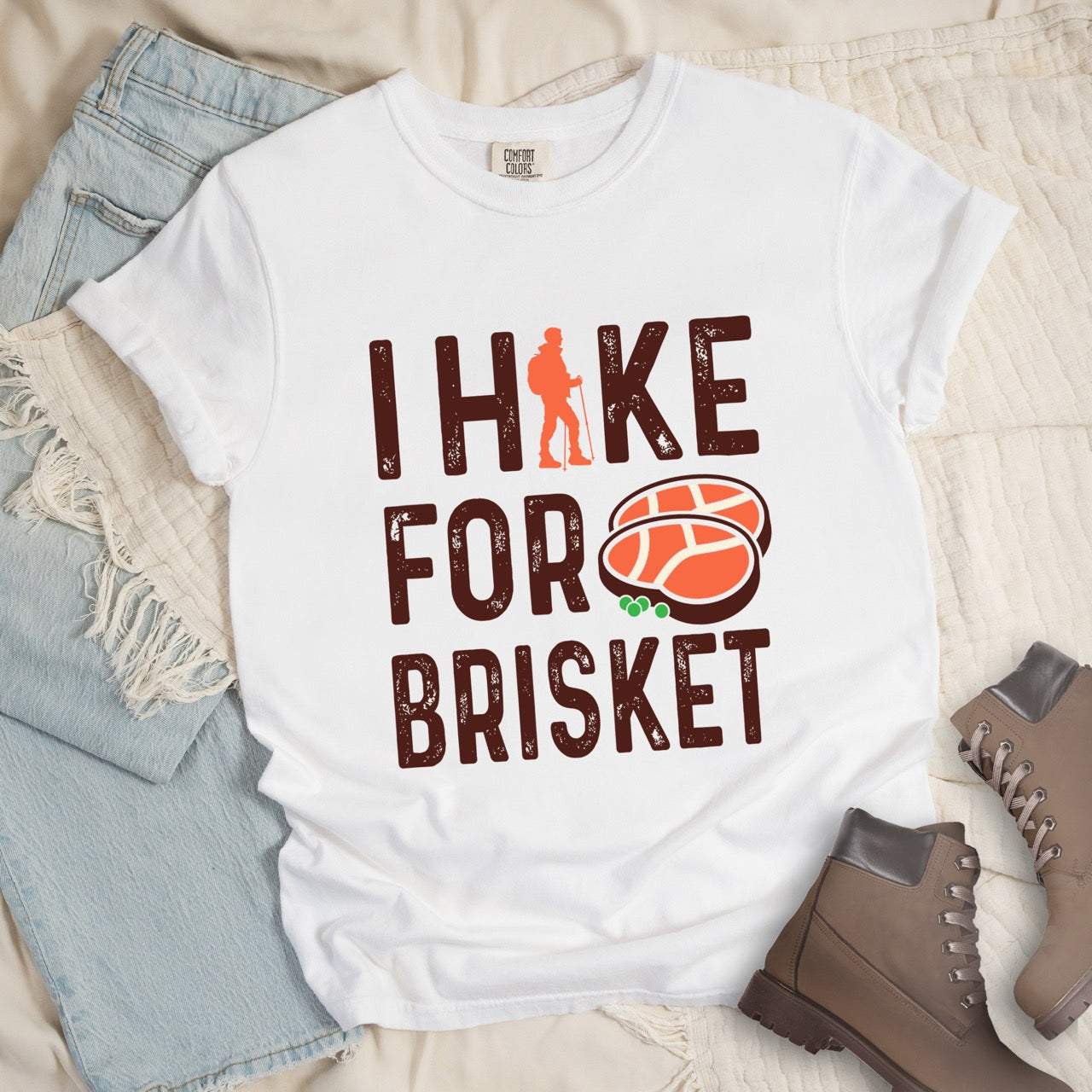 White BBQ brisket lover statement tee I Hike for Brisket with distressed brown typography character silhouette with trekking pole and illustrated sliced smoked brisket with herb garnish on vintage Comfort Colors shirt for food motivated adventurers for hikers and foodies - laid flat with pants and boots