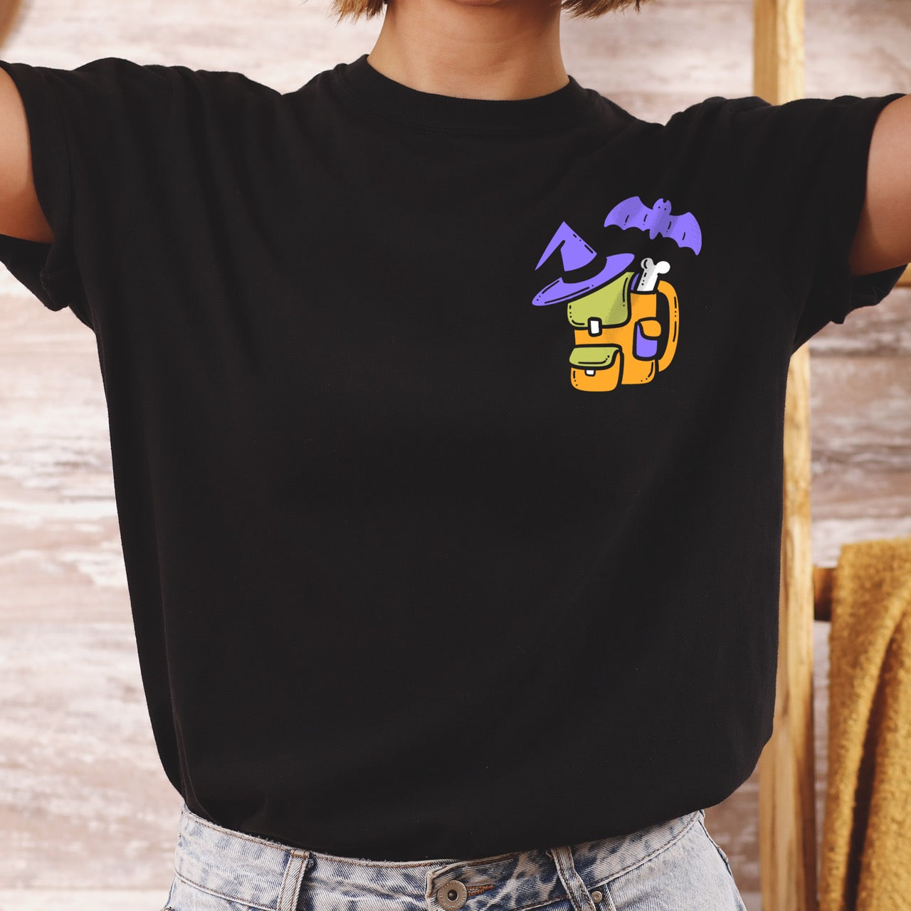 woman wearing Black t-shirt with colorful graphic design of a halloween themed back pack, witch hat and bat on a textured surface with jeans and boots.