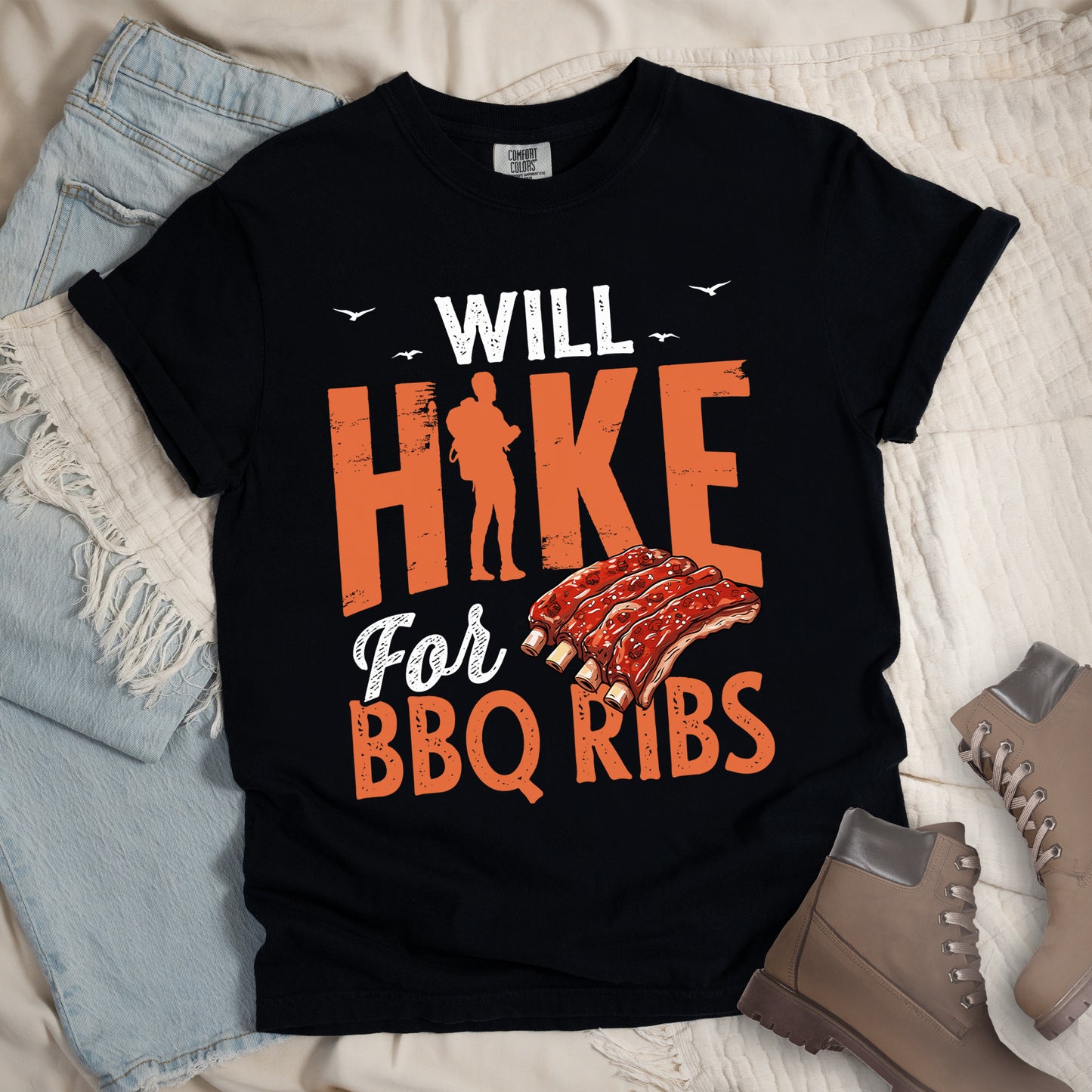 Black t-shirt with text "WILL HIKE For BBQ RIBS". "WILL" appears in white distressed letters at the top with small bird silhouettes. "HIKE" is in large orange distressed text with a hiker silhouette replacing the letter "I". "For" appears in white script, and "BBQ RIBS" in matching orange at the bottom. Between "For" and "BBQ RIBS" is an illustration of a rack of barbecue ribs with exposed bones. Features a classic crew neck and casual rolled sleeves, styled alongside light wash denim and brown hiking boots