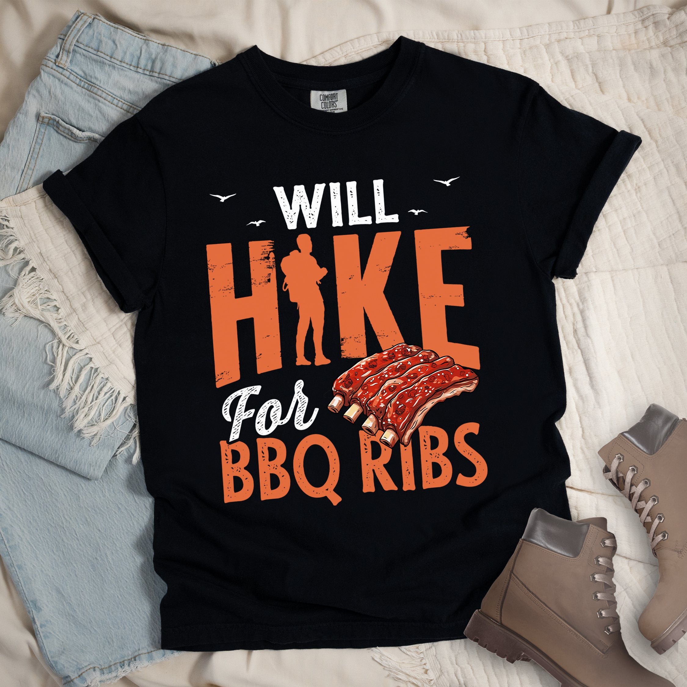 Black t-shirt with text "WILL HIKE For BBQ RIBS". "WILL" appears in deep brown-red distressed letters at the top with small bird silhouettes. "HIKE" is in large orange distressed text with a hiker silhouette replacing the letter "I". "For" appears in deep brown-red script, and "BBQ RIBS" in matching orange at the bottom. Between "For" and "BBQ RIBS" is an illustration of a rack of barbecue ribs with exposed bones.