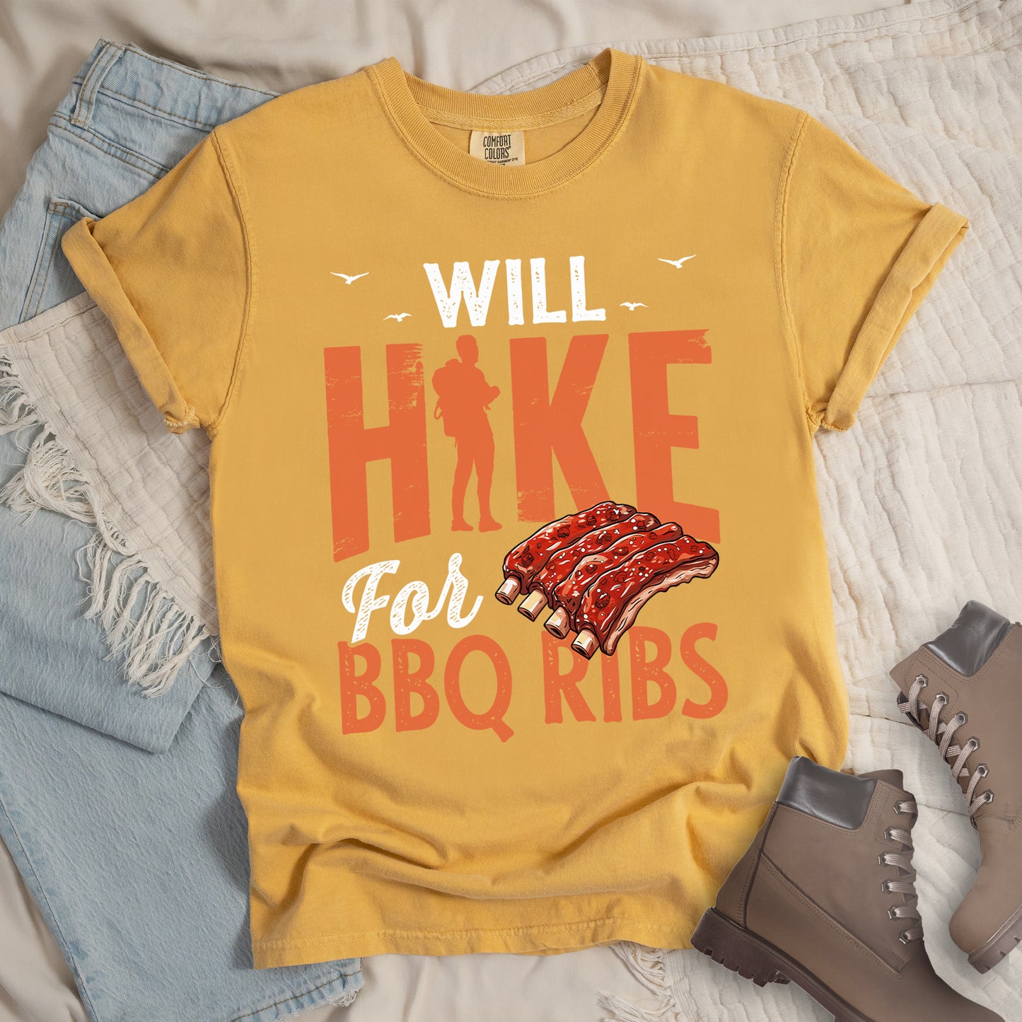Mustard (Yellow) t-shirt with text "WILL HIKE For BBQ RIBS". "WILL" appears in white distressed letters at the top with small bird silhouettes. "HIKE" is in large orange distressed text with a hiker silhouette replacing the letter "I". "For" appears in white script, and "BBQ RIBS" in matching orange at the bottom. Between "For" and "BBQ RIBS" is an illustration of a rack of barbecue ribs with exposed bones.