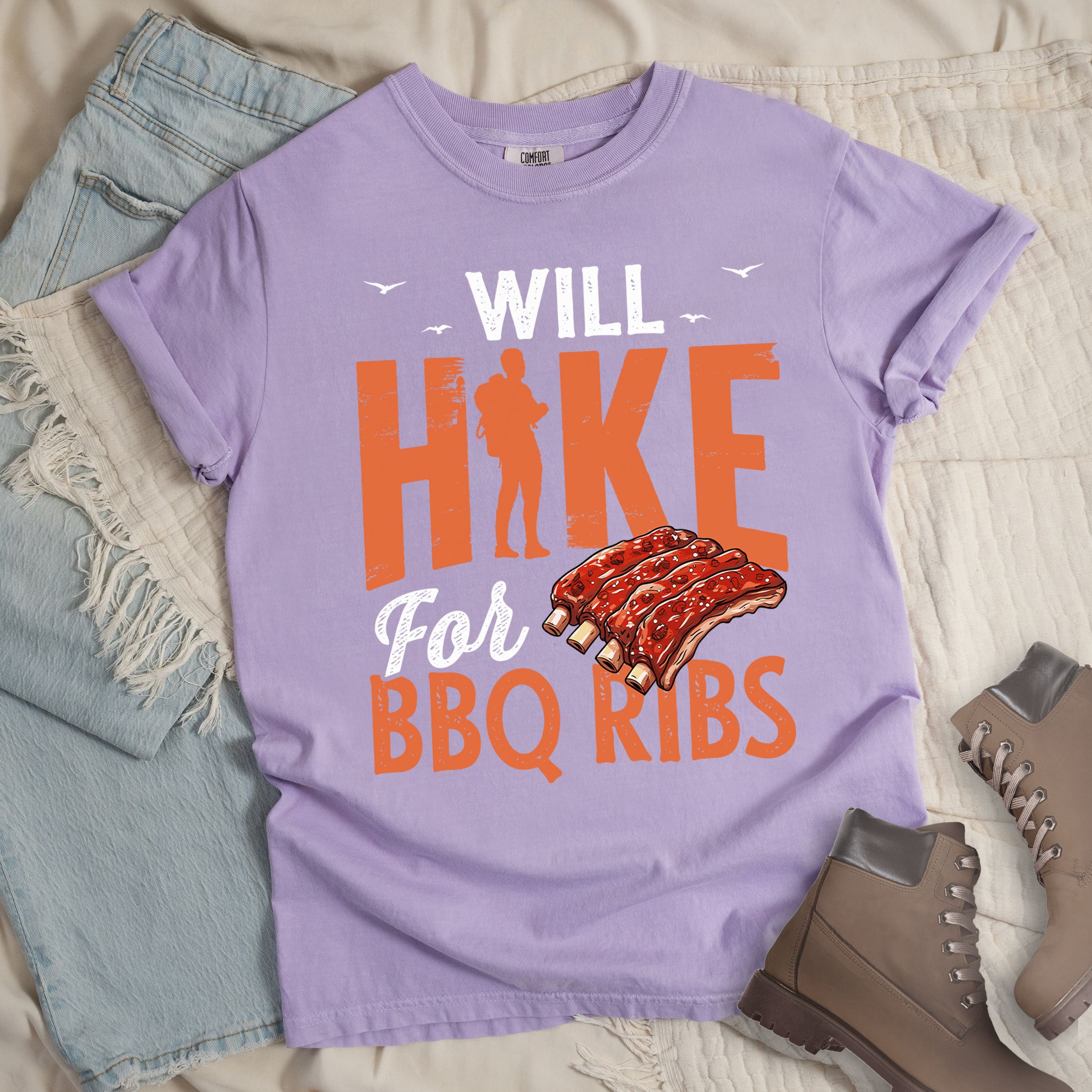 Light purple t-shirt with text "WILL HIKE For BBQ RIBS". "WILL" appears in deep brown-red distressed letters at the top with small bird silhouettes. "HIKE" is in large orange distressed text with a hiker silhouette replacing the letter "I". "For" appears in deep brown-red script, and "BBQ RIBS" in matching orange at the bottom. Between "For" and "BBQ RIBS" is an illustration of a rack of barbecue ribs with exposed bones.