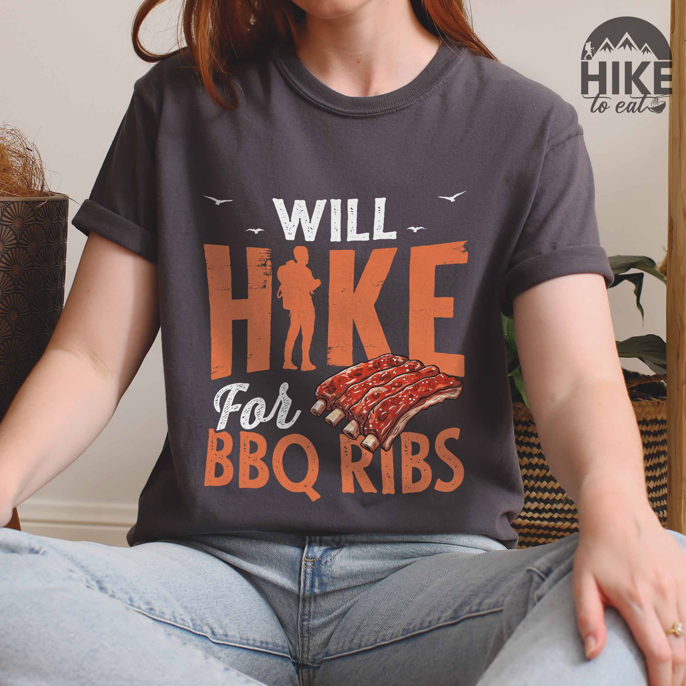Woman wearing pepper "Will Hike for BBQ Ribs" colored vintage tee with bold typography, birds, juicy grilled bbq ribs illustration, and backpacker for bbq enthusiasts