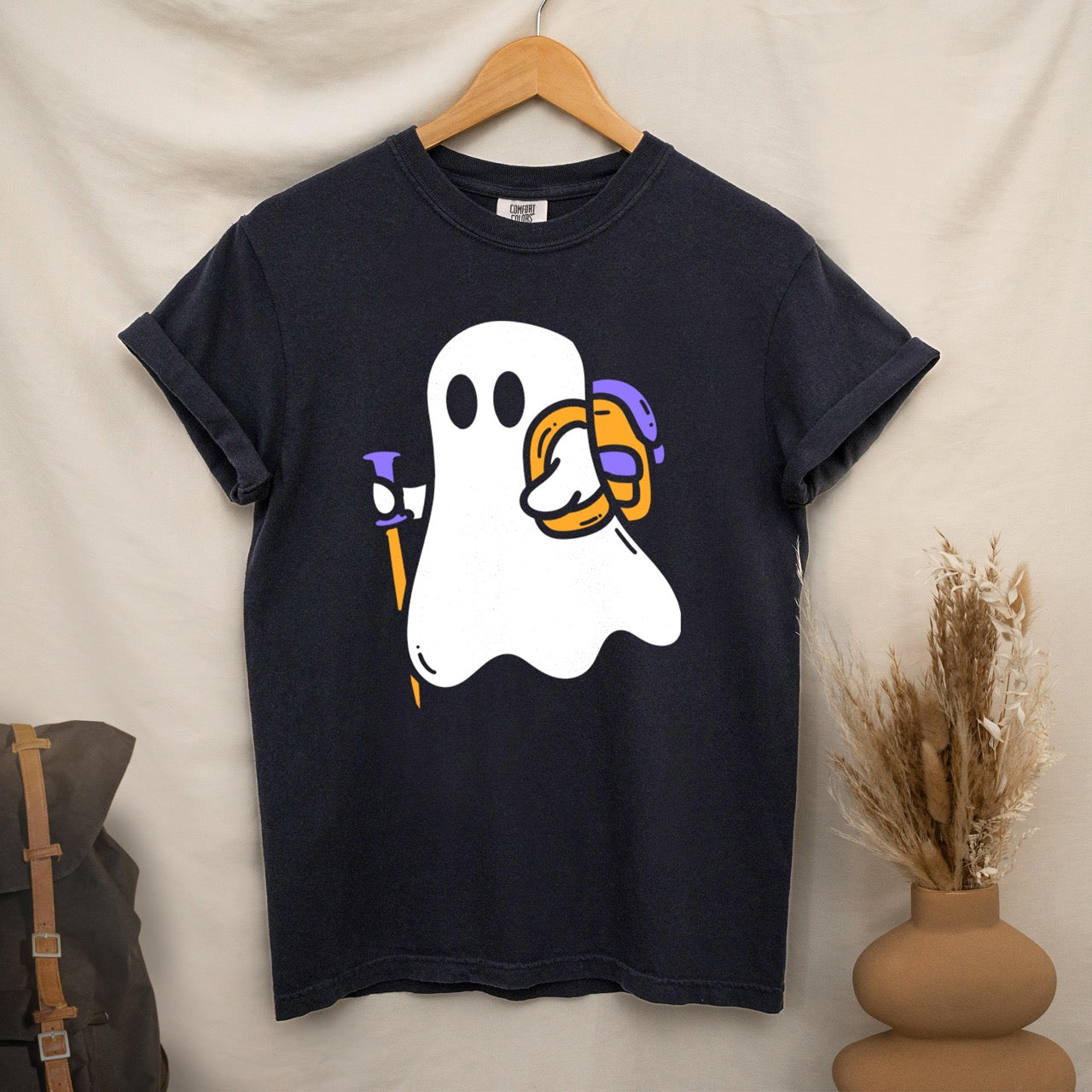 Hanging Black Halloween ghost statement tee with large full front design featuring cute white Boo character holding orange and purple trekking pole with colorful backpack perfect for spooky season celebrations on Comfort Colors shirt for hikers and adventurers.