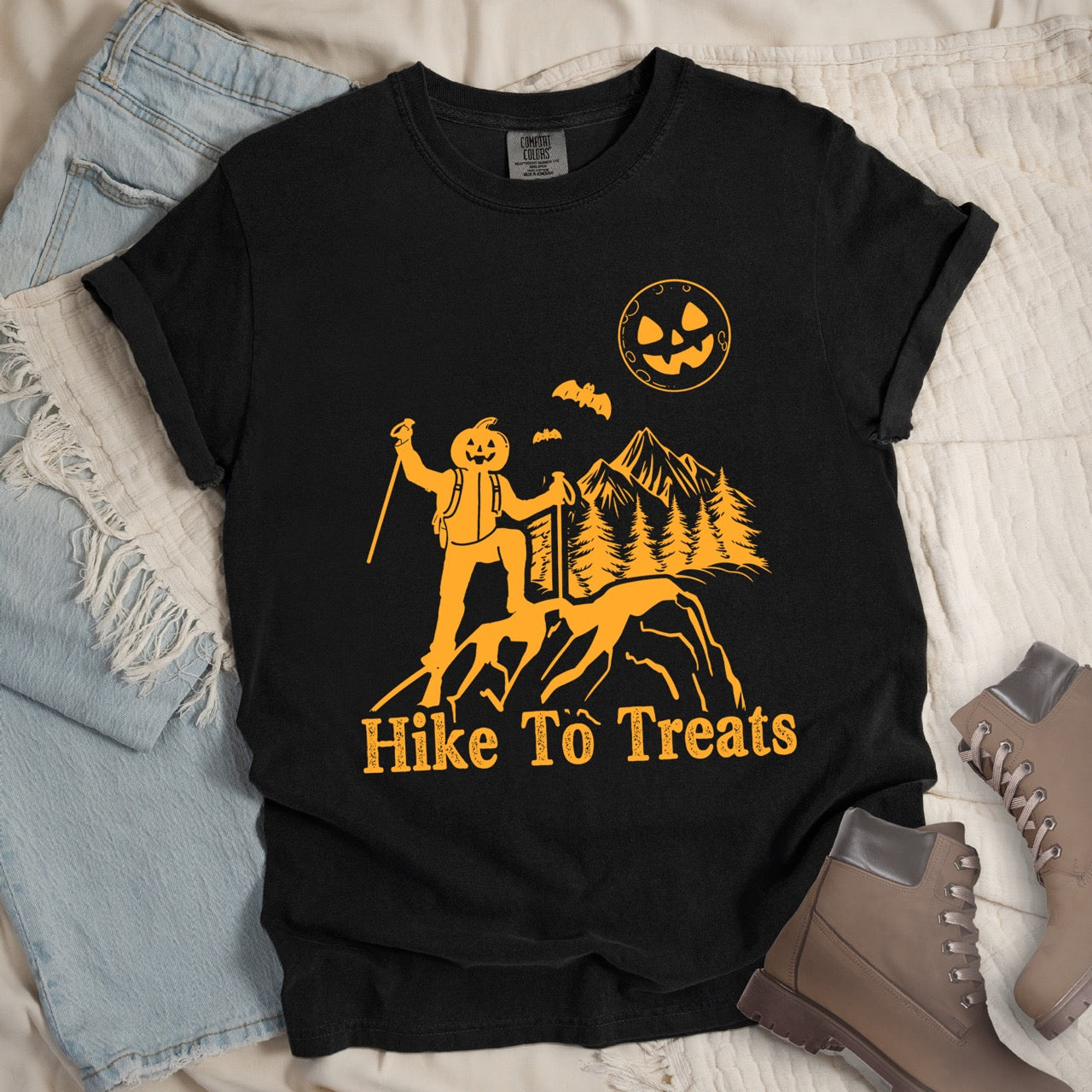 Black Limited edition Halloween statement shirt Hike To Treats featuring orange pumpkin head character silhouette with mountain landscape flying bats and jack-o-lantern moon on Comfort Colors tee for festive fall celebrations for hikers and foodies with shoes and pants.