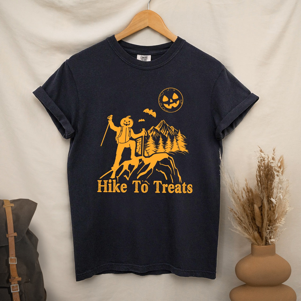 Black Limited edition Halloween statement shirt Hike To Treats featuring orange pumpkin head character silhouette with mountain landscape flying bats and jack-o-lantern moon on Comfort Colors tee for festive fall celebrations for hikers and foodies hanging on a wooden hanger.
