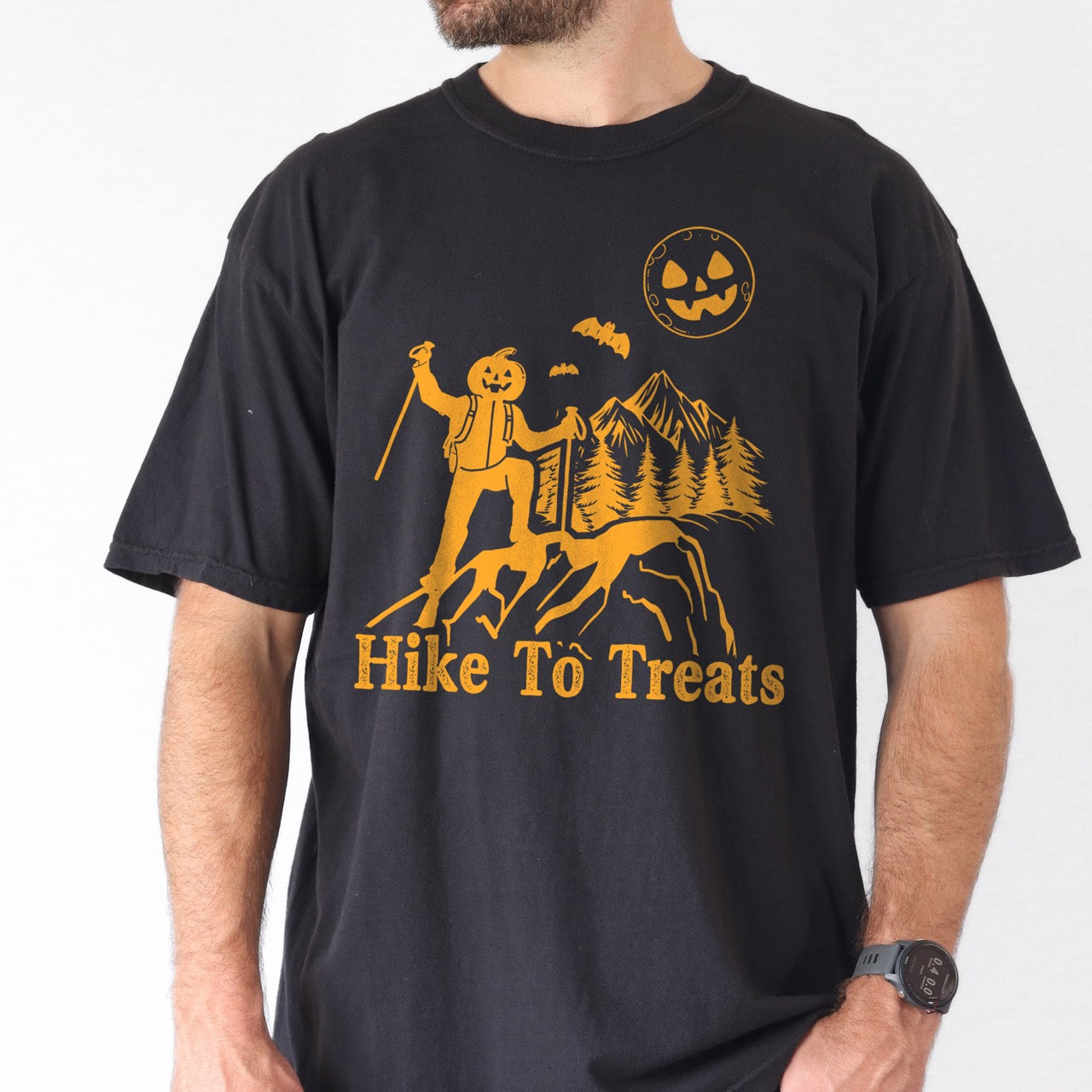 Man wearing Black Limited edition Halloween statement shirt Hike To Treats featuring orange pumpkin head character silhouette with mountain landscape flying bats and jack-o-lantern moon on Comfort Colors tee for festive fall celebrations for hikers and foodies.