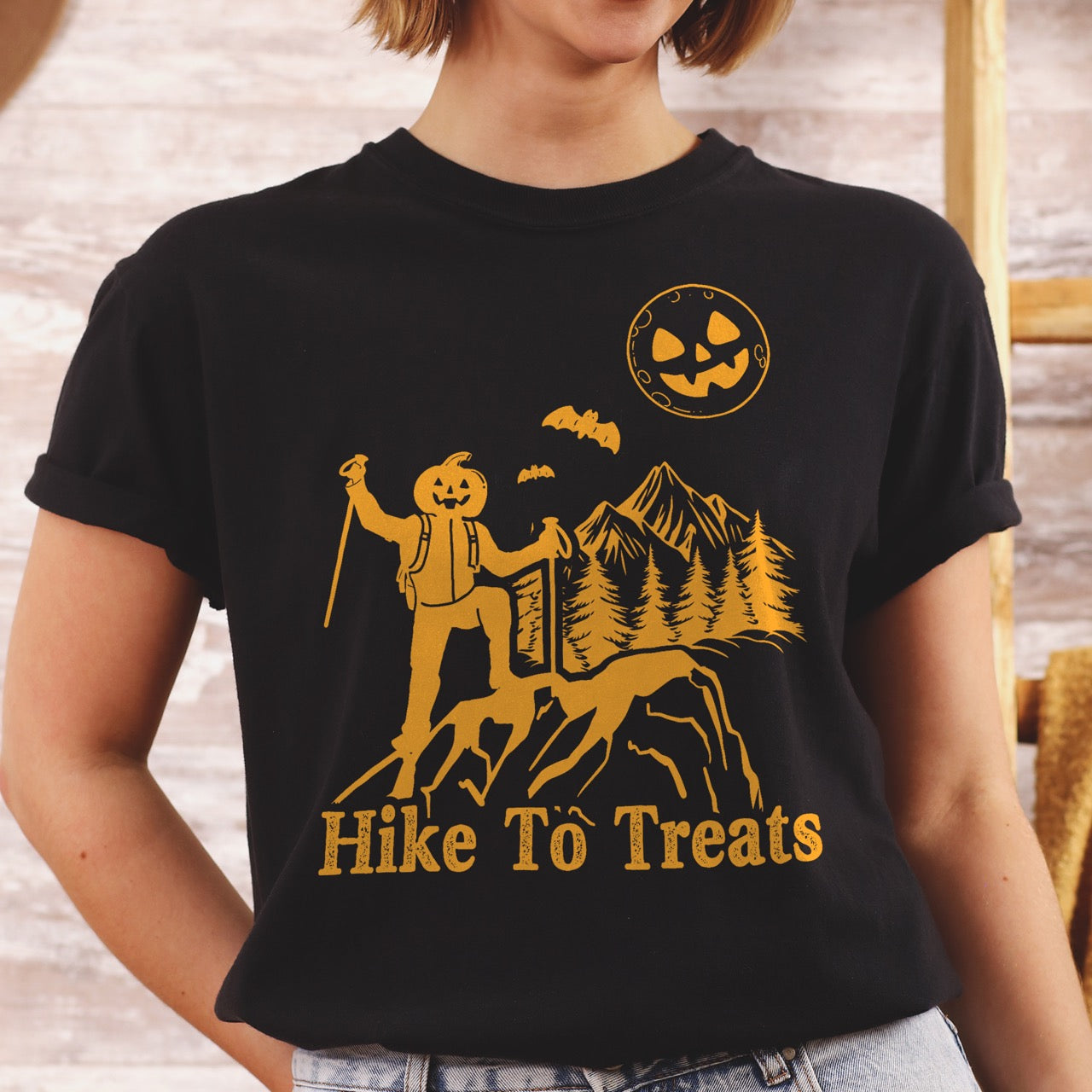 Woman wearing black Limited edition Halloween statement shirt Hike To Treats featuring orange pumpkin head character silhouette with mountain landscape flying bats and jack-o-lantern moon on Comfort Colors tee for festive fall celebrations for hikers and foodies.