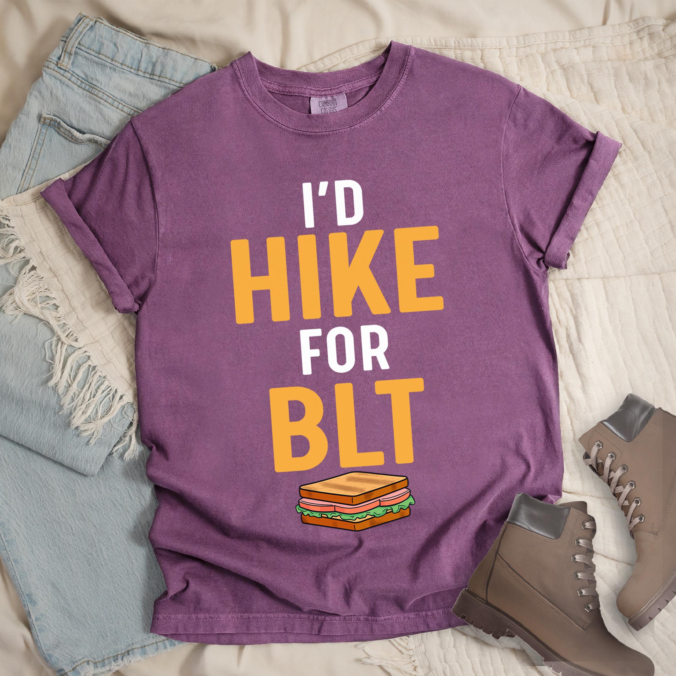Dark pink t-shirt with text "I'D HIKE FOR BLT". The words "I'D" appear in white at the top, "HIKE" in large gold/yellow letters in the middle, "FOR" in white, and "BLT" in matching gold/yellow at the bottom. Below the text is a colorful illustration of a BLT sandwich showing toast, maybe bacon, lettuce, and tomato layers. The simple, bold design emphasizes hiking motivation through food rewards.