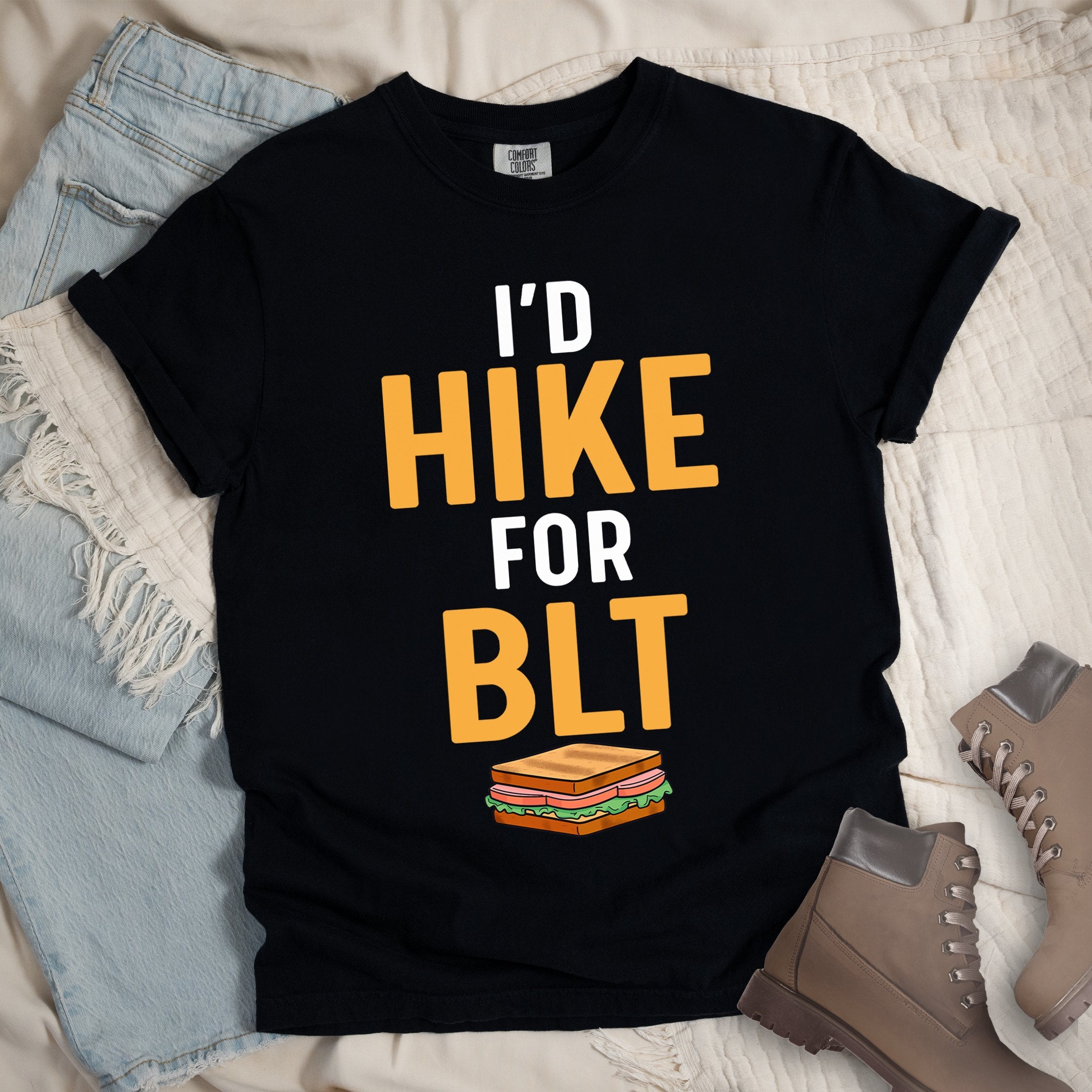 Black t-shirt with text "I'D HIKE FOR BLT". The words "I'D" appear in white at the top, "HIKE" in large gold/yellow letters in the middle, "FOR" in white, and "BLT" in matching gold/yellow at the bottom. Below the text is a colorful illustration of a BLT sandwich showing toast, maybe bacon, lettuce, and tomato layers. The simple, bold design emphasizes hiking motivation through food rewards.