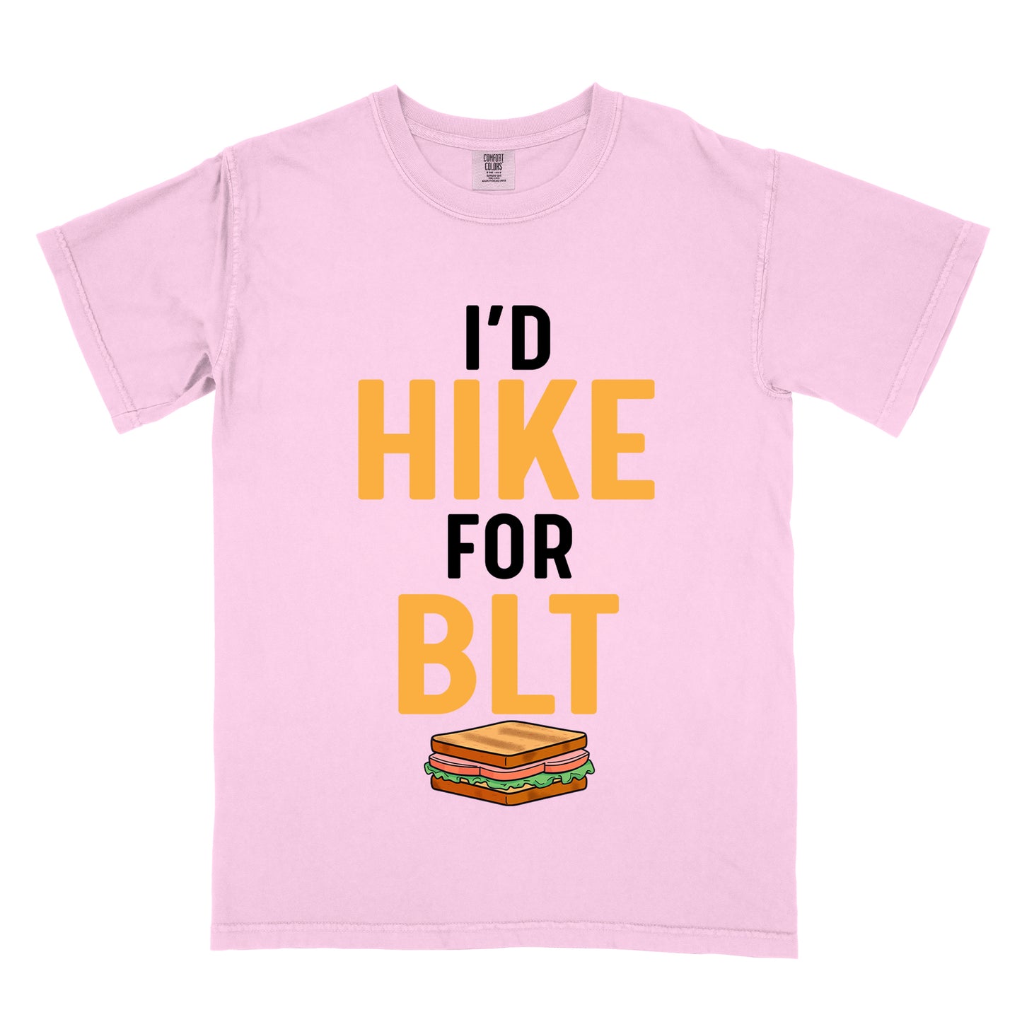 Blossom (Light Pink) t-shirt with text "I'D HIKE FOR BLT". The words "I'D" appear in black at the top, "HIKE" in large gold/yellow letters in the middle, "FOR" in black, and "BLT" in matching gold/yellow at the bottom. Below the text is a colorful illustration of a BLT sandwich showing toast, maybe bacon, lettuce, and tomato layers. The simple, bold design emphasizes hiking motivation through food rewards.