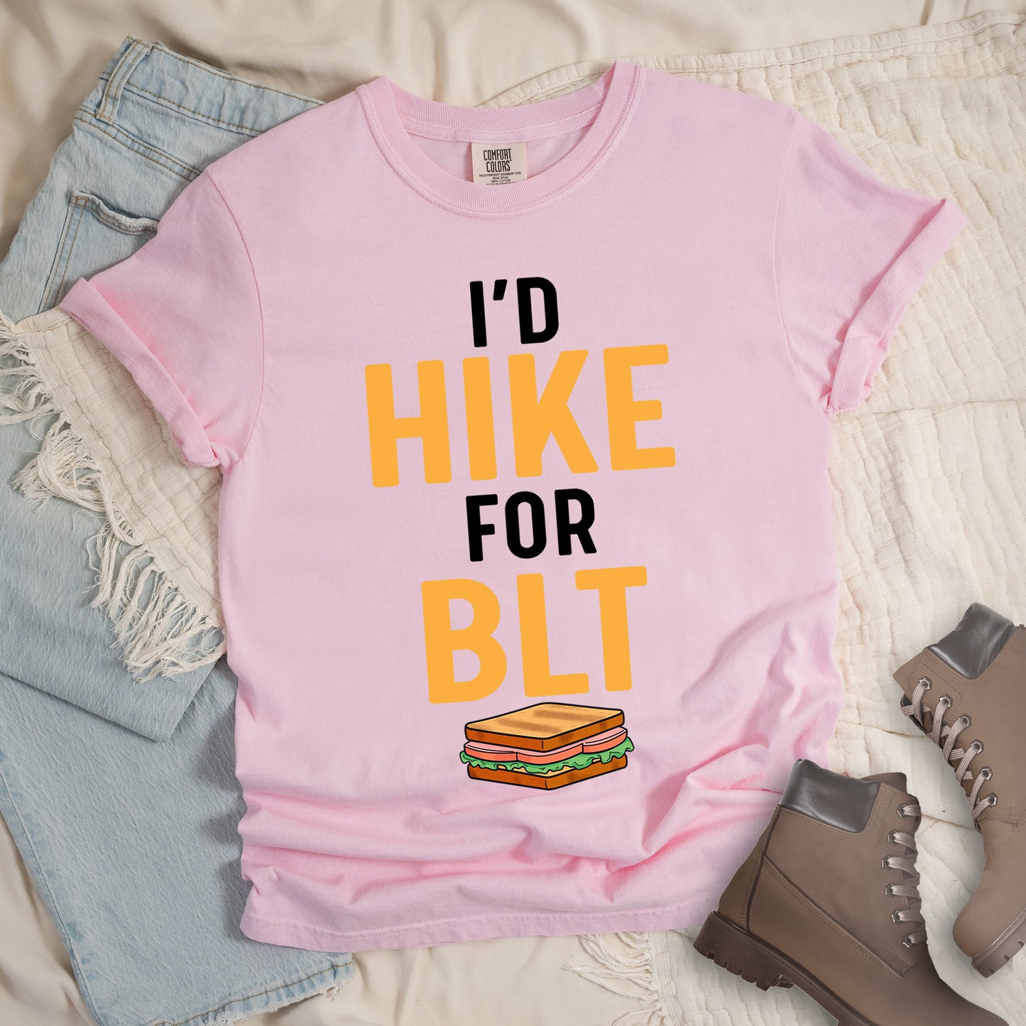Blossom (Light Pink) t-shirt with text "I'D HIKE FOR BLT". The words "I'D" appear in black at the top, "HIKE" in large gold/yellow letters in the middle, "FOR" in black, and "BLT" in matching gold/yellow at the bottom. Below the text is a colorful illustration of a BLT sandwich showing toast, maybe bacon, lettuce, and tomato layers. The simple, bold design emphasizes hiking motivation through food rewards.