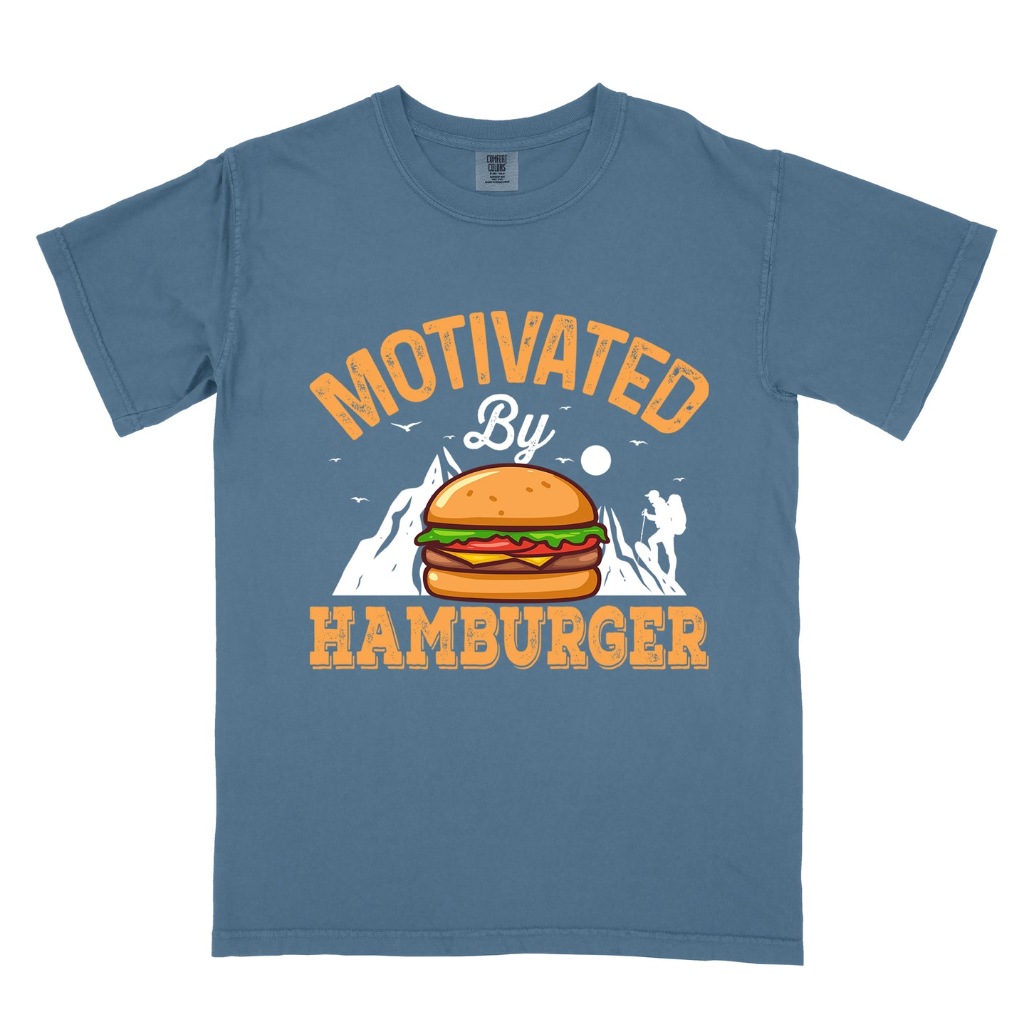 Blue Jean (Denim Blue) t-shirt with text "MOTIVATED By HAMBURGER". The word "MOTIVATED" appears in orange distressed letters at the top in an arc, "By" in white script in the middle, and "HAMBURGER" in matching orange at the bottom. The design features white mountain silhouettes with a small hiker figure, flying birds, and a sun or moon. A colorful hamburger illustration with bun, patty, cheese, lettuce and tomato sits in the center of the design.