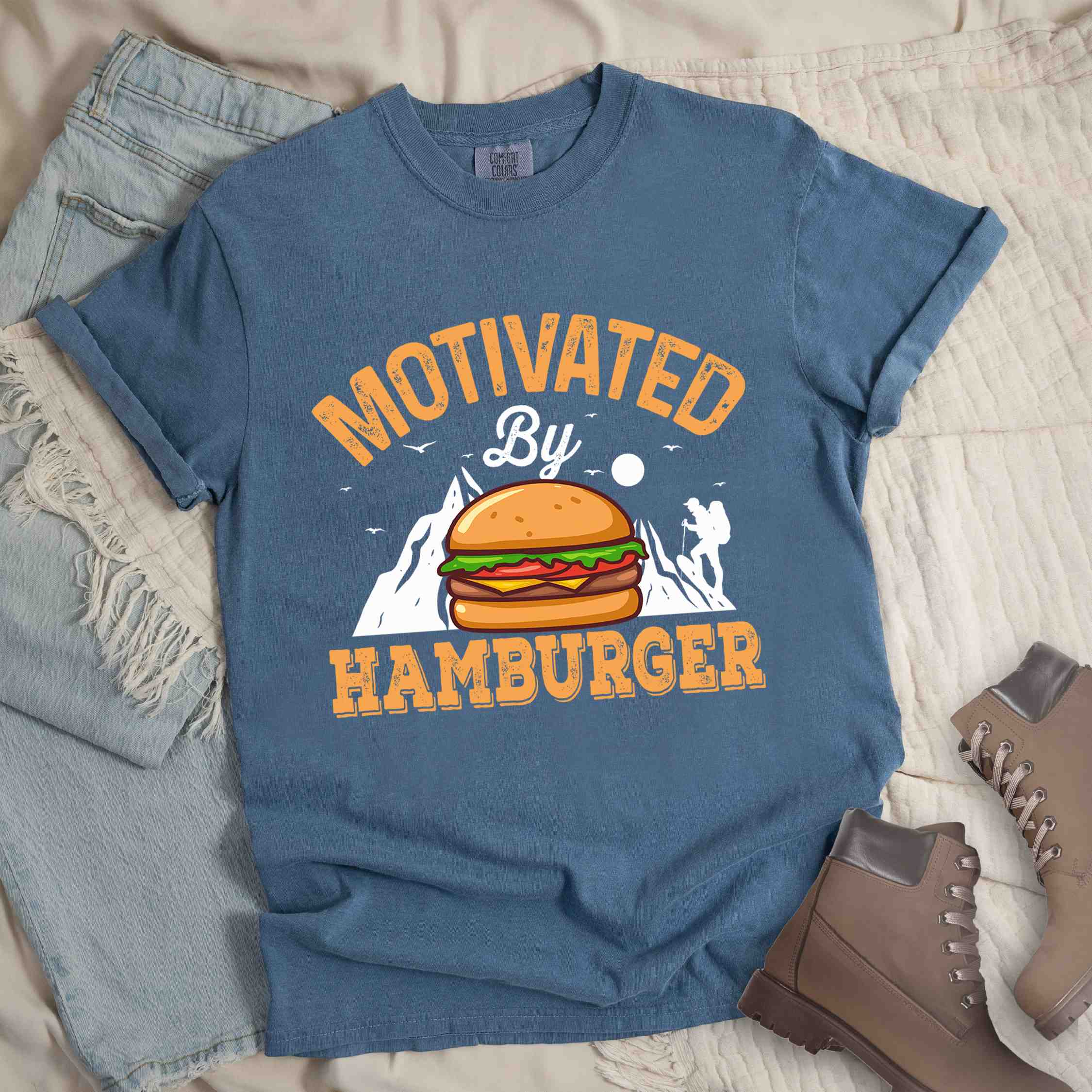 Blue jean "Motivated by Hamburger" colored vintage tee with bold typography, birds, mountains, illustrated juicy hamburger, and backpacker for burger enthusiasts.