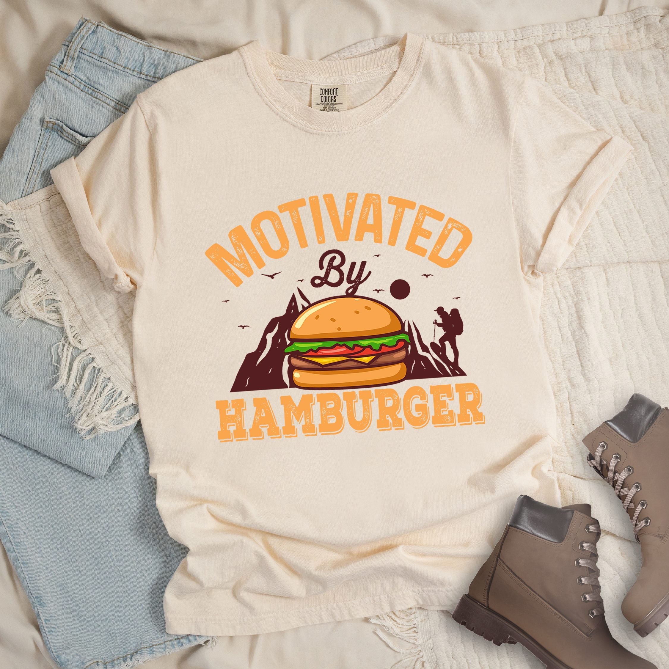 Ivory t-shirt with text "MOTIVATED By HAMBURGER". The word "MOTIVATED" appears in distressed letters at the top in an arc, "By" in script in the middle, and "HAMBURGER" in matching orange at the bottom. The design features white mountain silhouettes with a small hiker figure, flying birds, and a sun or moon. A colorful hamburger illustration with bun, patty, cheese, lettuce and tomato sits in the center of the design.