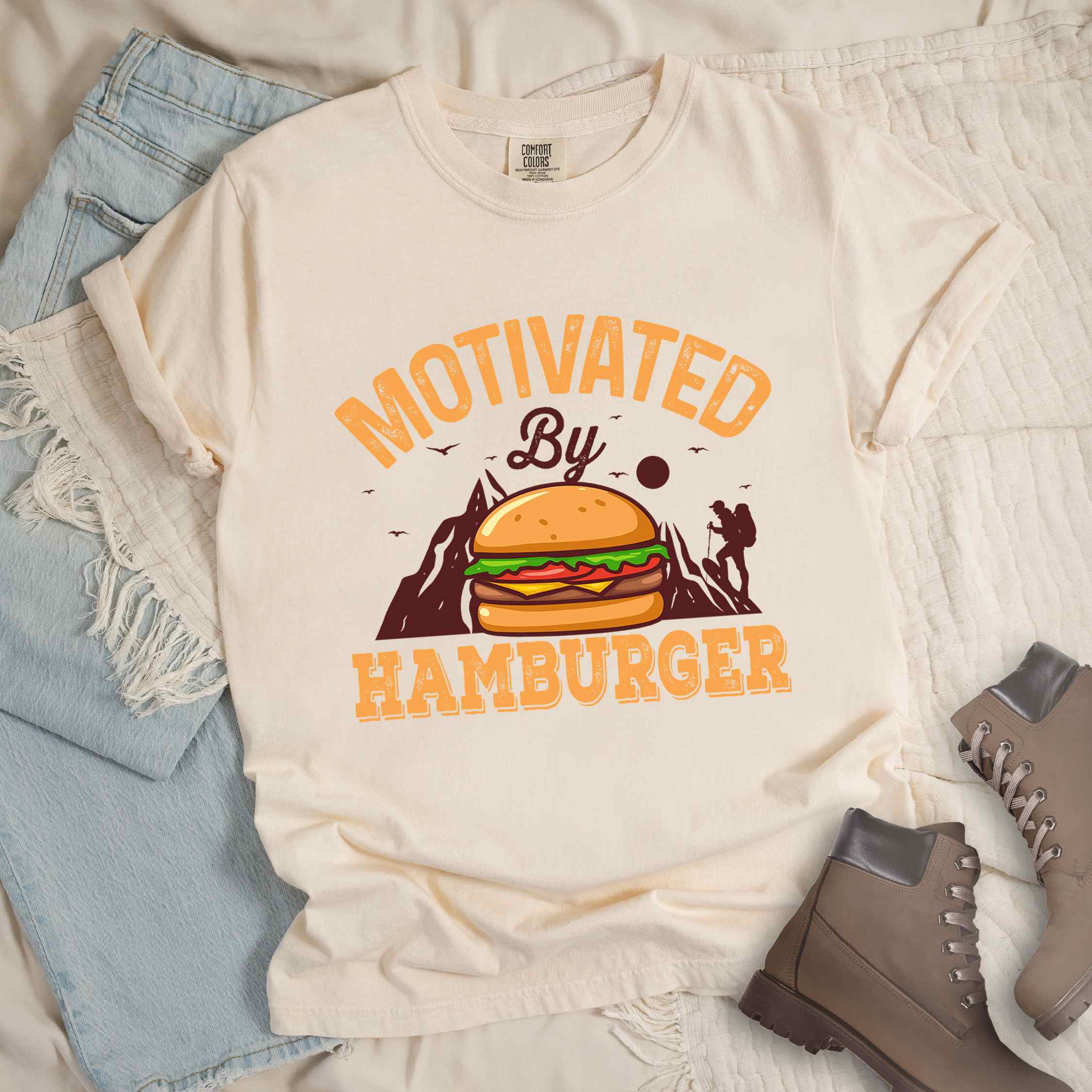 Ivory "Motivated by Hamburger" colored vintage tee with bold typography, birds, mountains, illustrated juicy hamburger, and backpacker for burger enthusiasts.