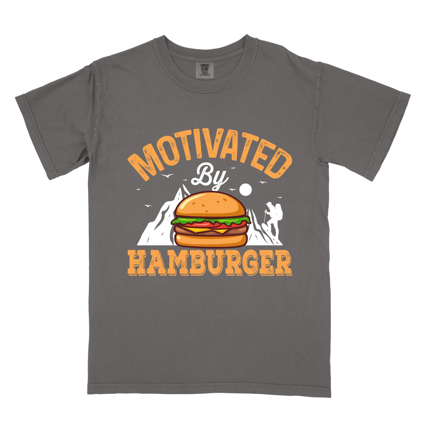 Pepper (Dark Gray) t-shirt with text "MOTIVATED By HAMBURGER". The word "MOTIVATED" appears in orange distressed letters at the top in an arc, "By" in white script in the middle, and "HAMBURGER" in matching orange at the bottom. The design features white mountain silhouettes with a small hiker figure, flying birds, and a sun or moon. A colorful hamburger illustration with bun, patty, cheese, lettuce and tomato sits in the center of the design.