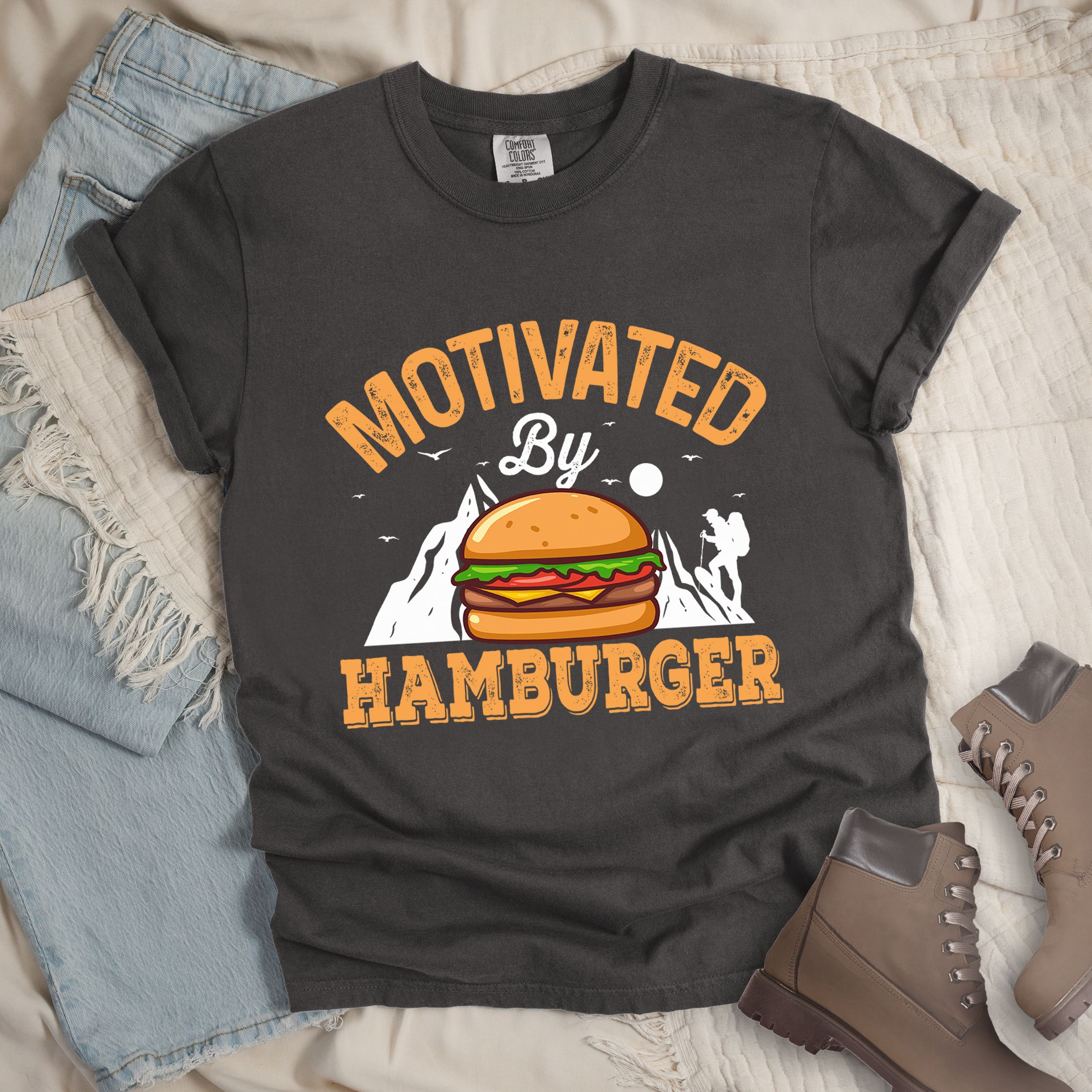 Dark grey t-shirt with text "MOTIVATED By HAMBURGER". The word "MOTIVATED" appears in distressed letters at the top in an arc, "By" in script in the middle, and "HAMBURGER" in matching orange at the bottom. The design features white mountain silhouettes with a small hiker figure, flying birds, and a sun or moon. A colorful hamburger illustration with bun, patty, cheese, lettuce and tomato sits in the center of the design.