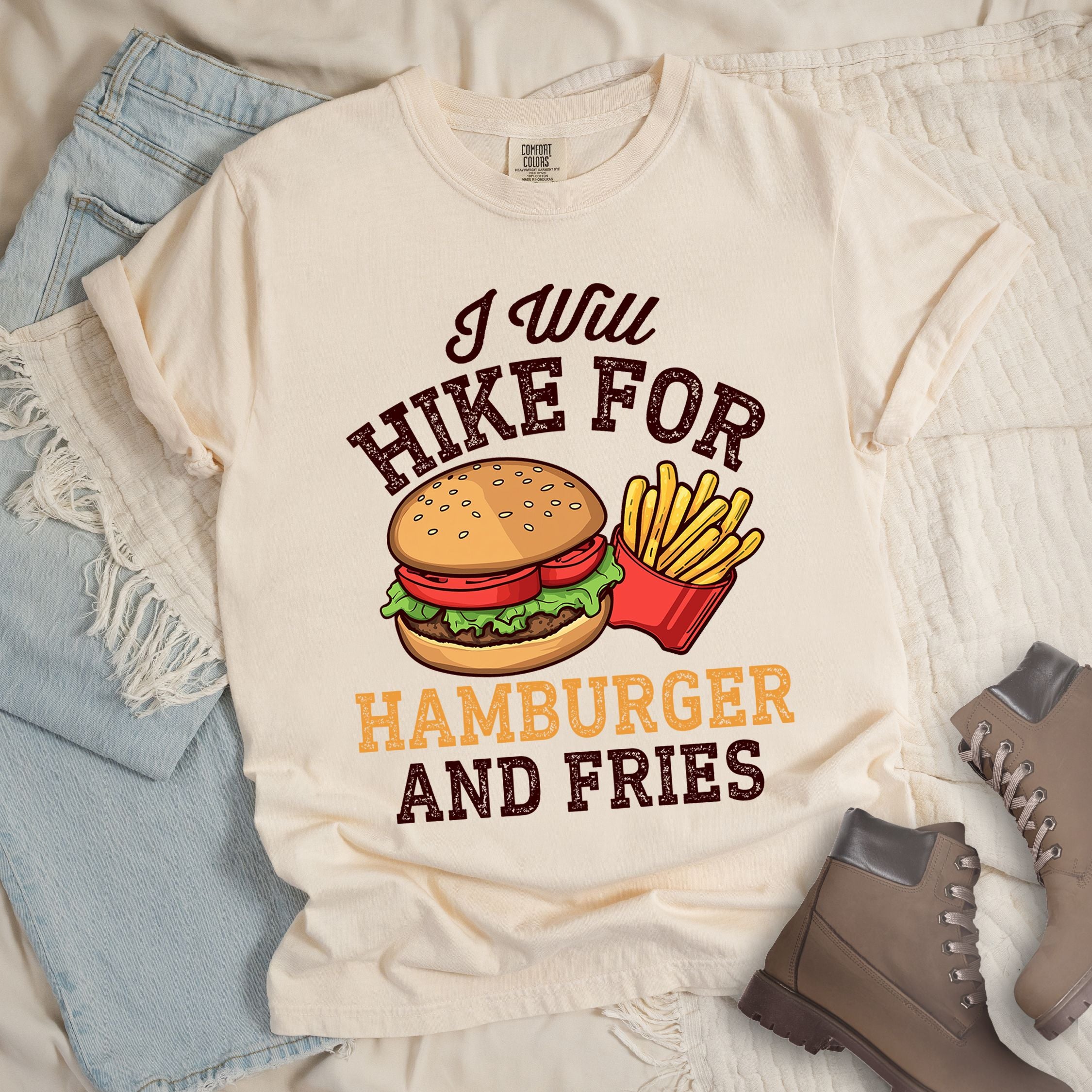 Ivory t-shirt with distressed text reading "I Will HIKE FOR HAMBURGER AND FRIES". The words "I Will" appear in script at the top, "HIKE FOR" in white block letters, "HAMBURGER" in gold/orange, and "AND FRIES" in white at the bottom. Center illustration shows a detailed hamburger with bun, patty, lettuce, and tomato next to a red container of yellow french fries.