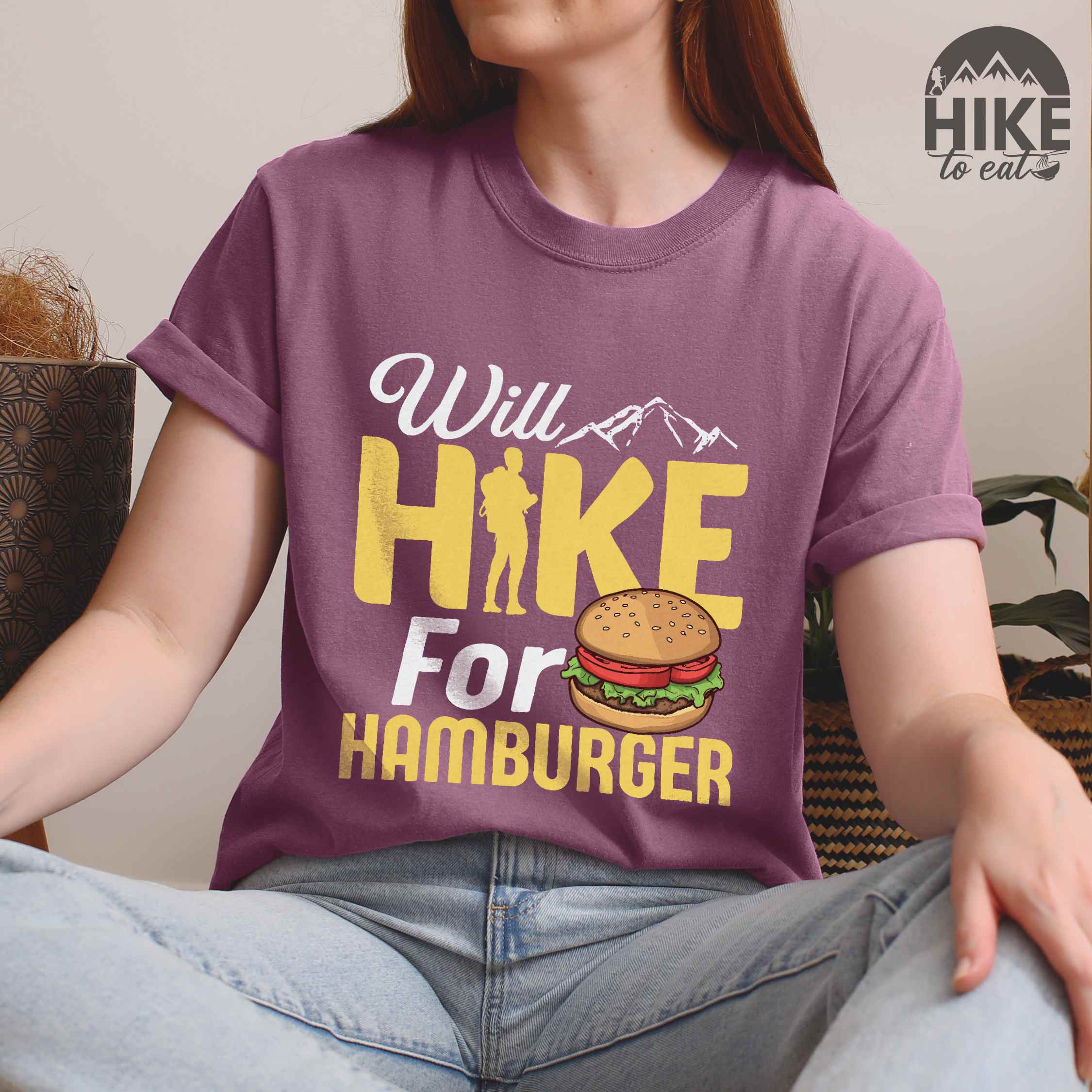 Woman wearing berry "Will Hike for Hamburger" colored vintage tee with bold typography, birds, mountains, illustrated juicy hamburger, and backpacker for burger enthusiasts.
