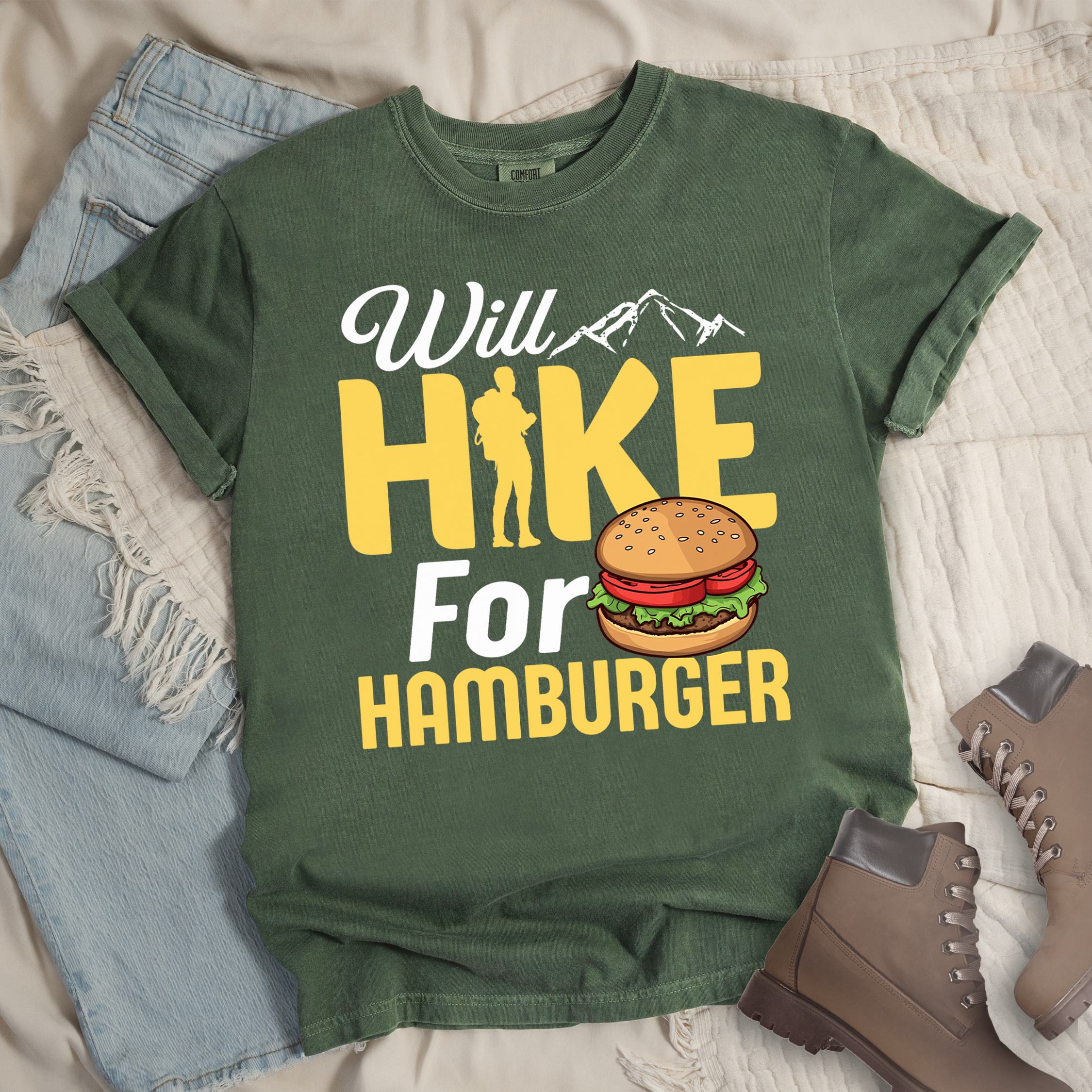 Dark green t-shirt with text "Will HIKE For HAMBURGER". "Will" appears in white script with small mountain silhouettes. "HIKE" is in large yellow letters with a hiker silhouette replacing the letter "I". "For" text, and "HAMBURGER" at the bottom. Next to "For" and above "HAMBURGER" is a colorful illustration of a hamburger with sesame seed bun, lettuce, tomato, and patty.Features a classic crew neck and casual rolled sleeves, styled alongside light wash denim and brown hiking boots