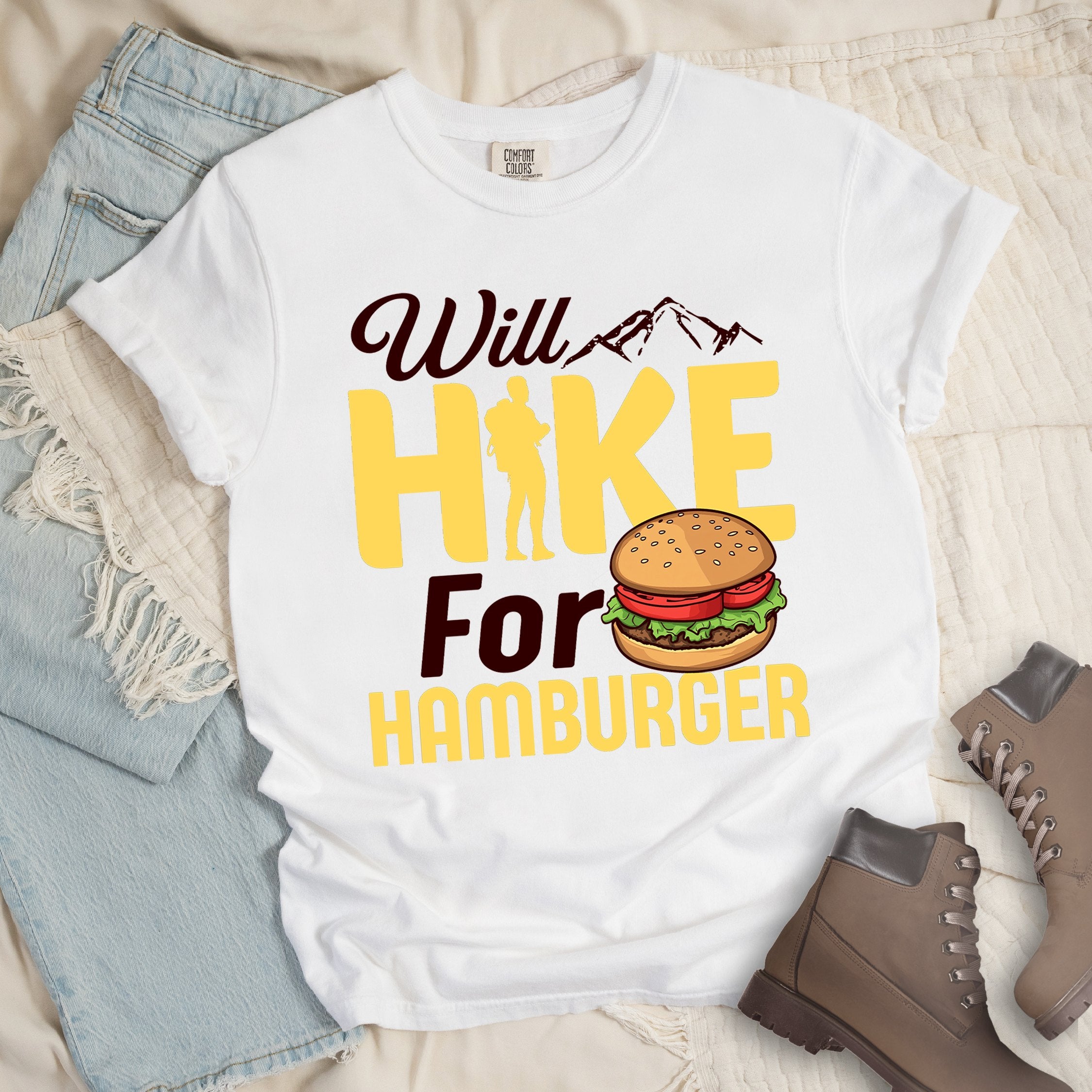White t-shirt with text "Will HIKE For HAMBURGER". "Will" appears in white script with small mountain silhouettes. "HIKE" is in large yellow letters with a hiker silhouette replacing the letter "I". "For" text, and "HAMBURGER" at the bottom. Next to "For" and above "HAMBURGER" is a colorful illustration of a hamburger with sesame seed bun, lettuce, tomato, and patty.Features a classic crew neck and casual rolled sleeves, styled alongside light wash denim and brown hiking boots