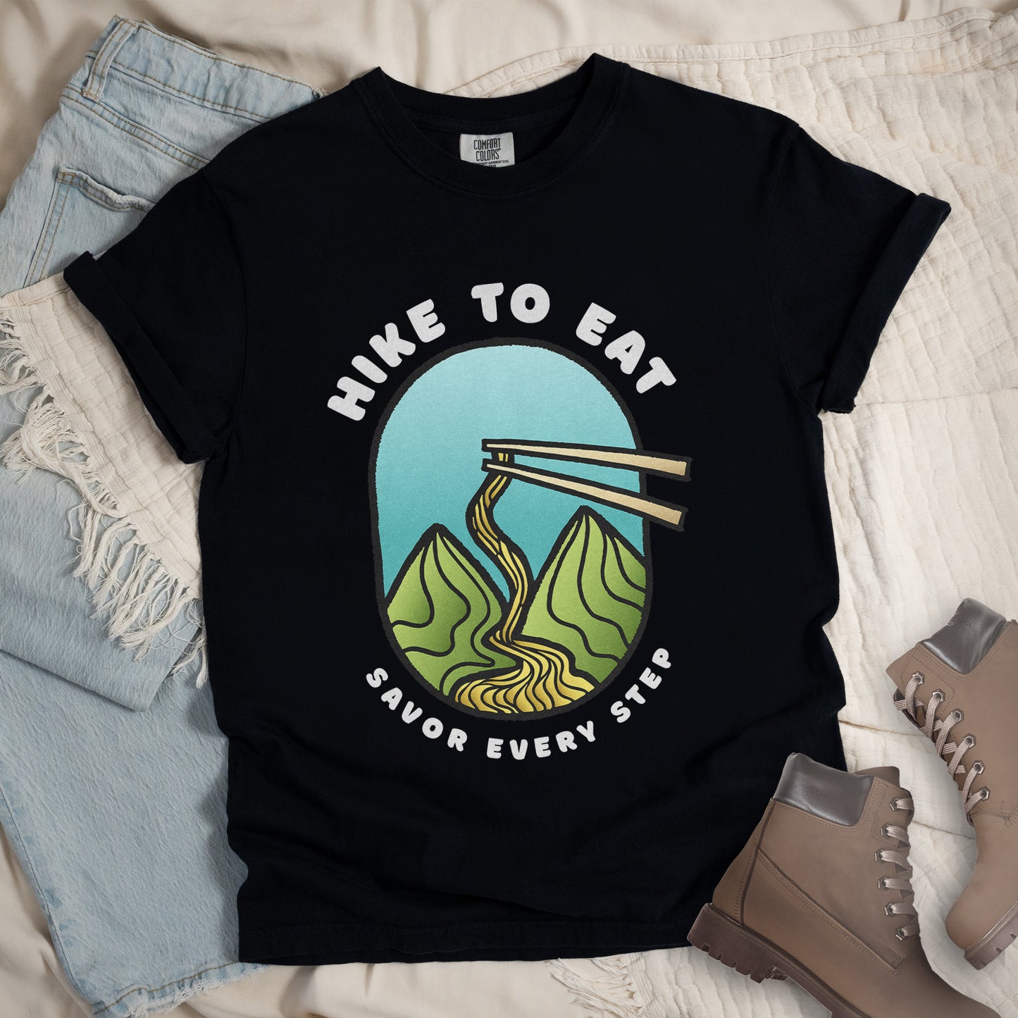 A black t-shirt with white text "HIKE TO EAT" arched at the top and "SAVOR EVERY STEP" at the bottom. The center features an oval graphic with a turquoise blue sky background, green mountain peaks, and chopsticks lifting ramen noodles that flow like a trail between the mountains. The shirt features a classic crew neck and casual rolled sleeves, styled alongside light wash denim and brown hiking boots.