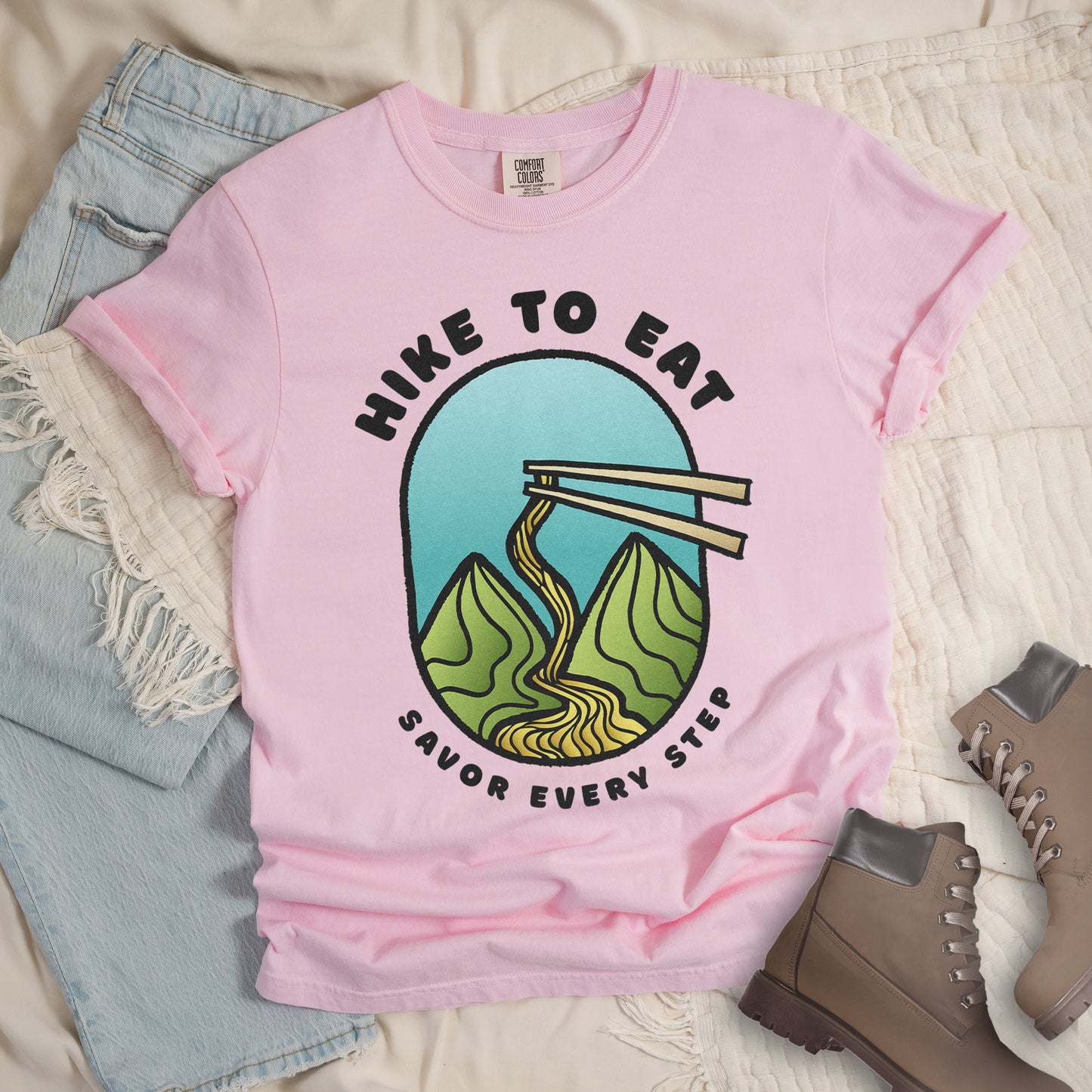 A blossom(light pink) t-shirt with black text "HIKE TO EAT" arched at the top and "SAVOR EVERY STEP" at the bottom. The center features an oval graphic with a turquoise blue sky background, green mountain peaks, and chopsticks lifting ramen noodles that flow like a trail between the mountains. The shirt features a classic crew neck and casual rolled sleeves, styled alongside light wash denim and brown hiking boots.