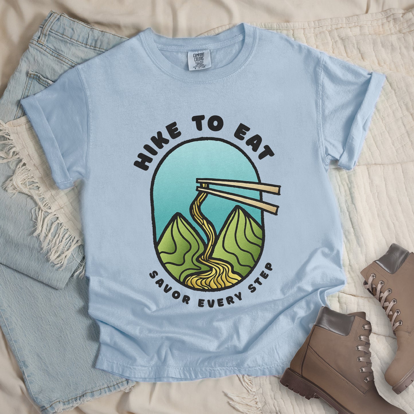 A chambray (light blue) t-shirt with black text "HIKE TO EAT" arched at the top and "SAVOR EVERY STEP" at the bottom. The center features an oval graphic with a turquoise blue sky background, green mountain peaks, and chopsticks lifting ramen noodles that flow like a trail between the mountains. The shirt features a classic crew neck and casual rolled sleeves, styled alongside light wash denim and brown hiking boots.