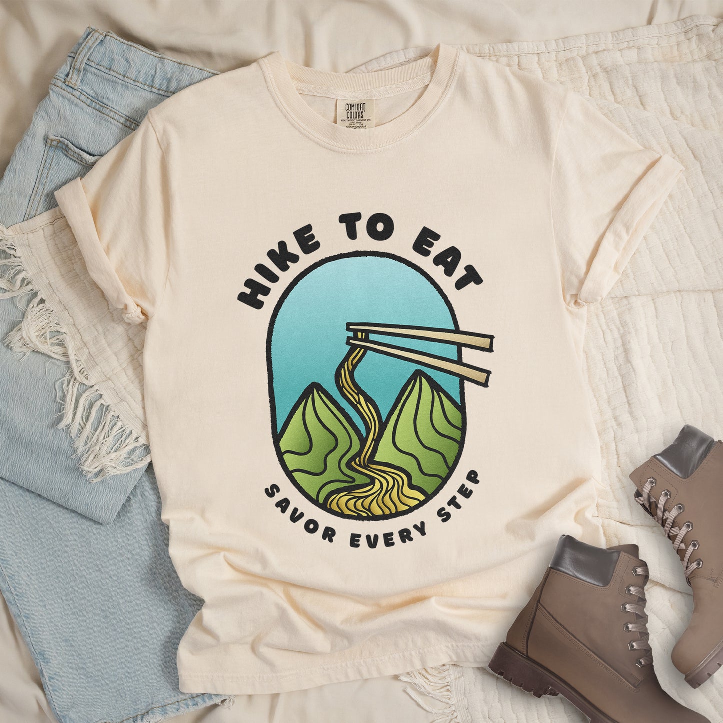 An ivory (cream white) t-shirt with black text "HIKE TO EAT" arched at the top and "SAVOR EVERY STEP" at the bottom. The center features an oval graphic with a turquoise blue sky background, green mountain peaks, and chopsticks lifting ramen noodles that flow like a trail between the mountains. The shirt features a classic crew neck and casual rolled sleeves, styled alongside light wash denim and brown hiking boots.