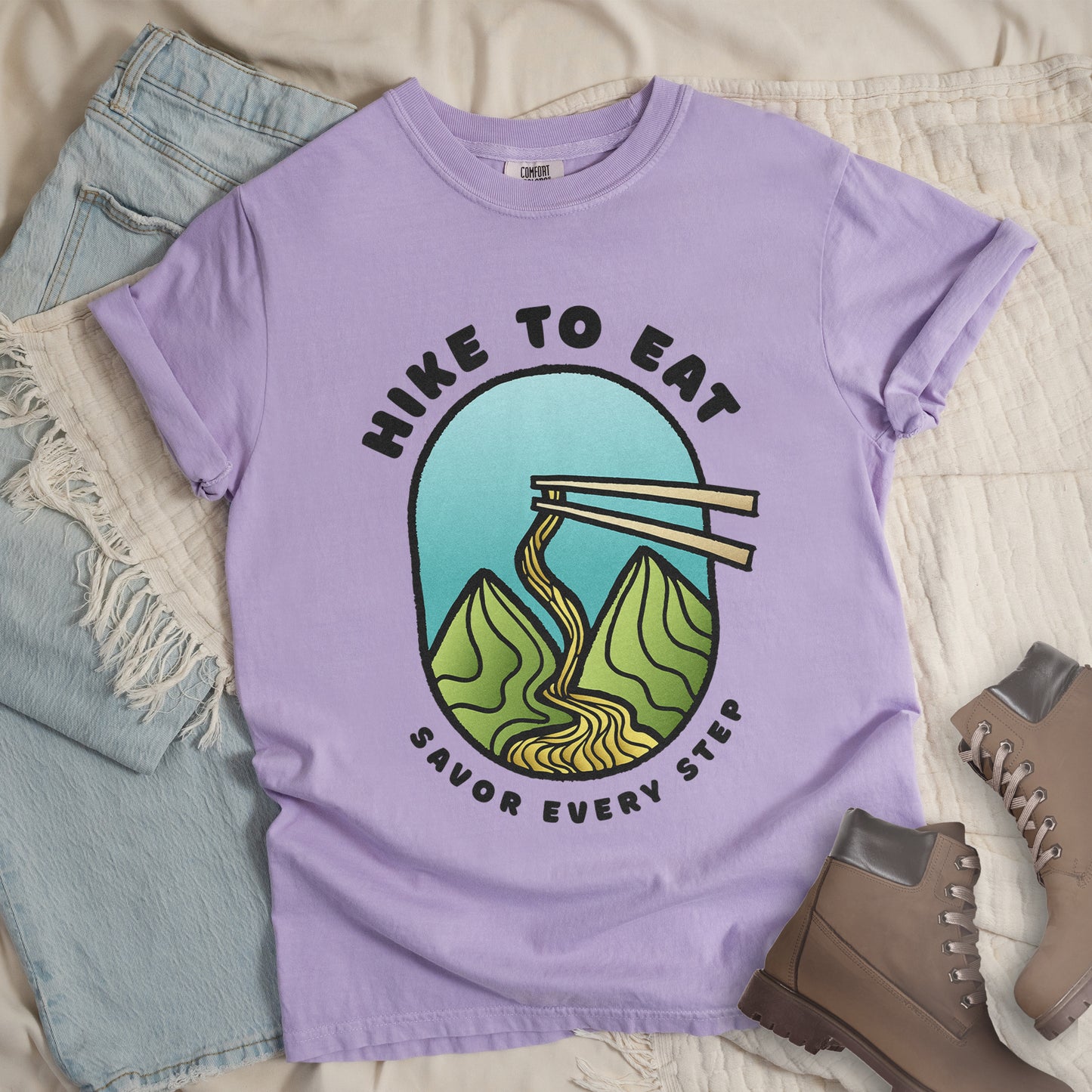 An orchid (light purple) t-shirt with black text "HIKE TO EAT" arched at the top and "SAVOR EVERY STEP" at the bottom. The center features an oval graphic with a turquoise blue sky background, green mountain peaks, and chopsticks lifting ramen noodles that flow like a trail between the mountains. The shirt features a classic crew neck and casual rolled sleeves, styled alongside light wash denim and brown hiking boots.