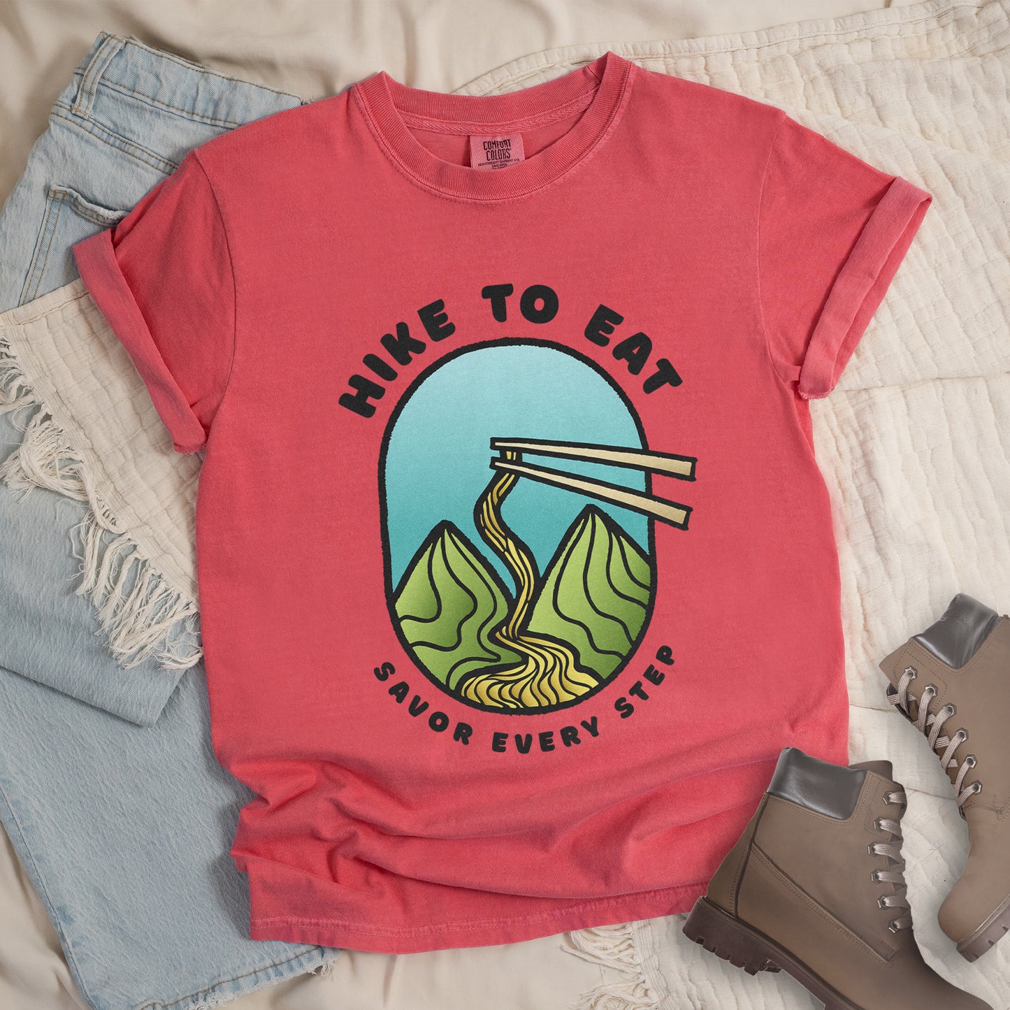 A watermelon (light coral) t-shirt with black text "HIKE TO EAT" arched at the top and "SAVOR EVERY STEP" at the bottom. The center features an oval graphic with a turquoise blue sky background, green mountain peaks, and chopsticks lifting ramen noodles that flow like a trail between the mountains. The shirt features a classic crew neck and casual rolled sleeves, styled alongside light wash denim and brown hiking boots.