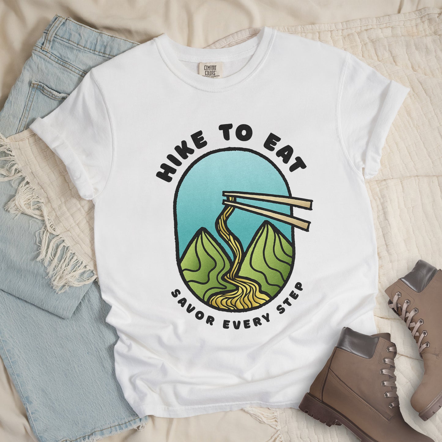 A white t-shirt with black text "HIKE TO EAT" arched at the top and "SAVOR EVERY STEP" at the bottom. The center features an oval graphic with a turquoise blue sky background, green mountain peaks, and chopsticks lifting ramen noodles that flow like a trail between the mountains. The shirt features a classic crew neck and casual rolled sleeves, styled alongside light wash denim and brown hiking boots.