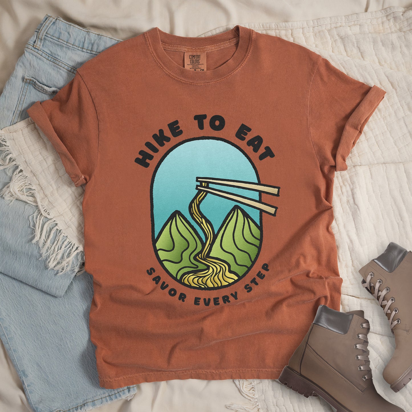 A yam (brown) t-shirt with black text "HIKE TO EAT" arched at the top and "SAVOR EVERY STEP" at the bottom. The center features an oval graphic with a turquoise blue sky background, green mountain peaks, and chopsticks lifting ramen noodles that flow like a trail between the mountains. The shirt features a classic crew neck and casual rolled sleeves, styled alongside light wash denim and brown hiking boots.