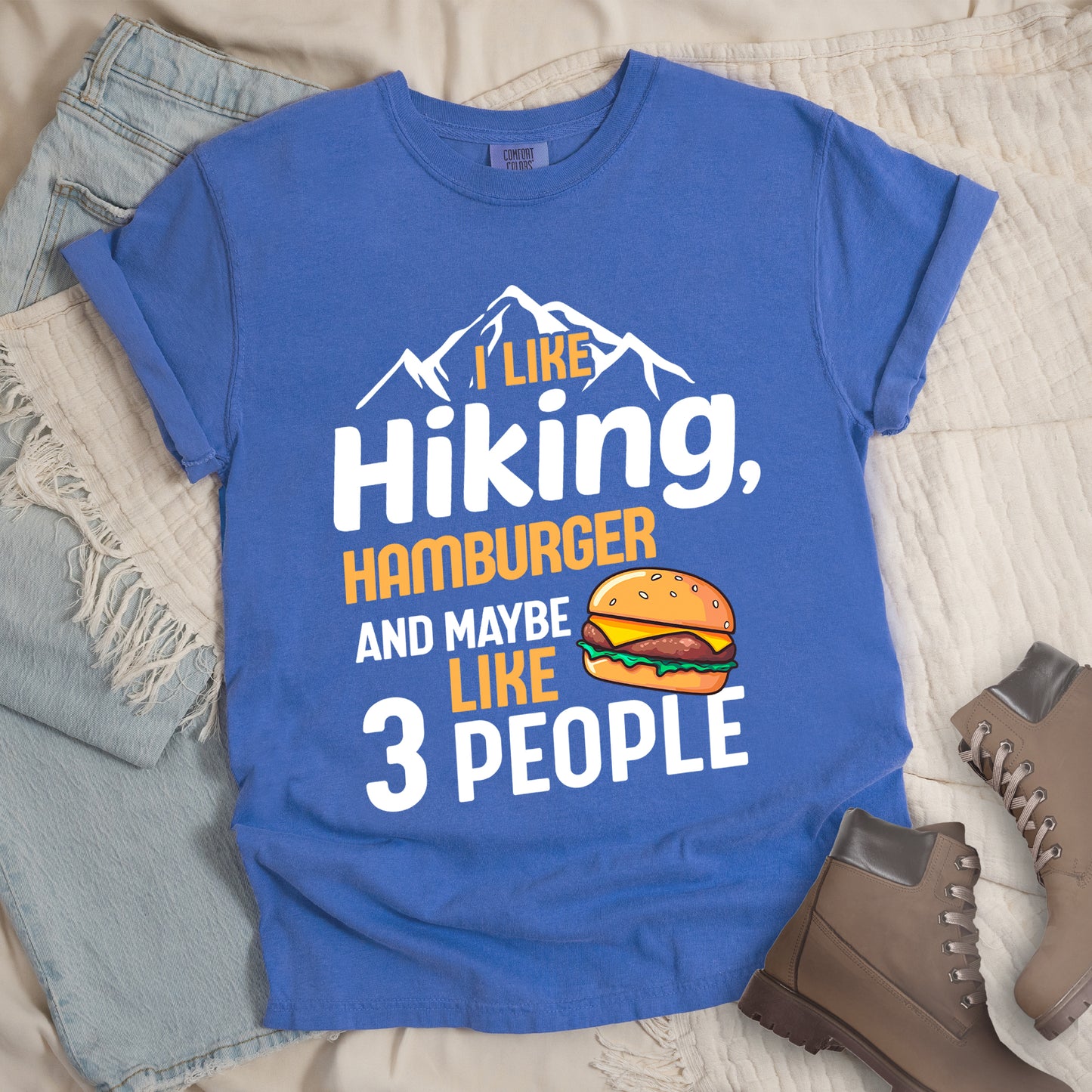 Flo Blue (Bright Blue) t-shirt with text "I LIKE Hiking, HAMBURGER AND MAYBE LIKE 3 PEOPLE". White mountain silhouettes appear at the top. "I LIKE" in orange, "Hiking," in white, "HAMBURGER" in orange, "AND MAYBE" in white, "LIKE" in orange, and "3 PEOPLE" in white. An illustration of a hamburger with sesame seed bun, cheese, lettuce and patty appears next to the word "LIKE". The design combines hiking and introvert humor.