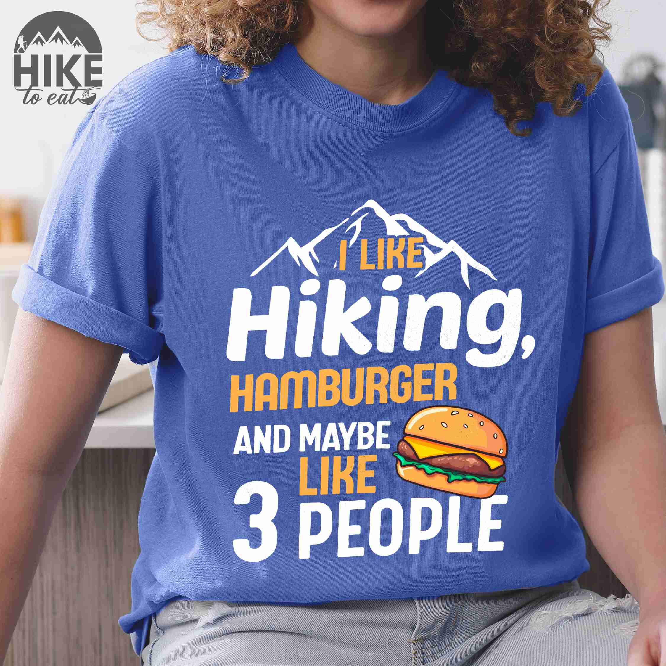 Woman wearing flo blue "I like hiking, hamburger and maybe like three people" colored vintage tee with bold typography, birds, mountains, illustrated juicy hamburger, and backpacker for burger enthusiasts.