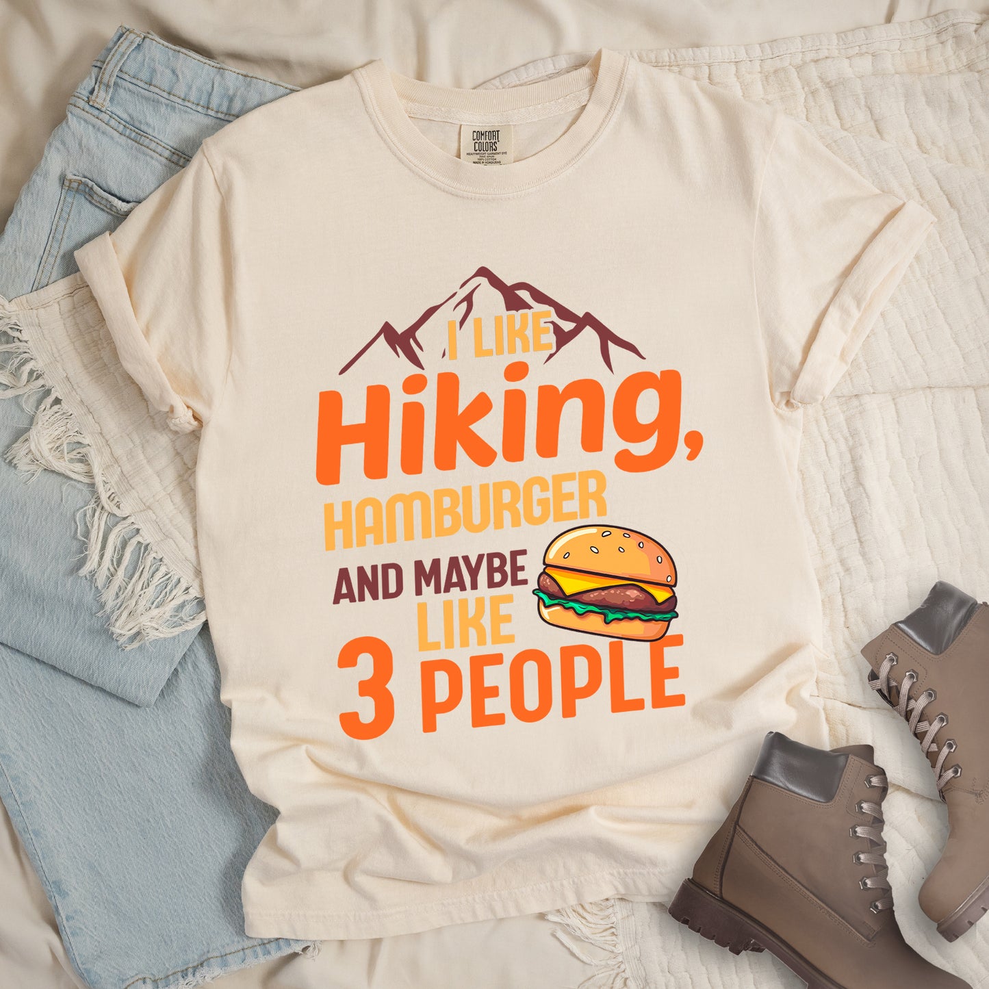 Ivory (Cream White) t-shirt with text "I LIKE Hiking, HAMBURGER AND MAYBE LIKE 3 PEOPLE". Maroon mountain silhouettes appear at the top. "I LIKE" in yellow, "Hiking," in orange, "HAMBURGER" in yellow, "AND MAYBE" in maroon, "LIKE" in yellow, and "3 PEOPLE" in orange. An illustration of a hamburger with sesame seed bun, cheese, lettuce and patty appears next to the word "LIKE". The design combines hiking and introvert humor.