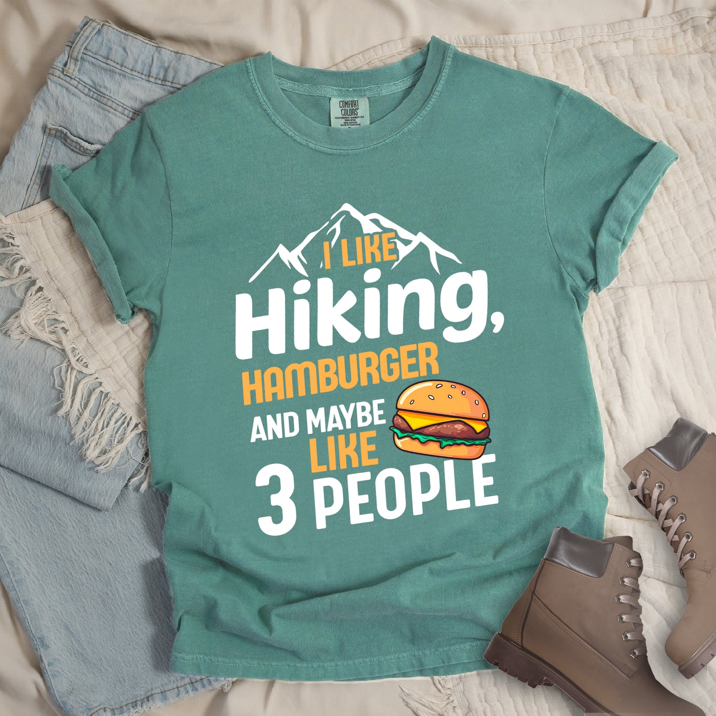 Light Green t-shirt with text "I LIKE Hiking, HAMBURGER AND MAYBE LIKE 3 PEOPLE". White mountain silhouettes appear at the top. "I LIKE" in orange, "Hiking," in white, "HAMBURGER" in orange, "AND MAYBE" in white, "LIKE" in orange, and "3 PEOPLE" in white. An illustration of a hamburger with sesame seed bun, cheese, lettuce and patty appears next to the word "LIKE". The design combines hiking and introvert humor.