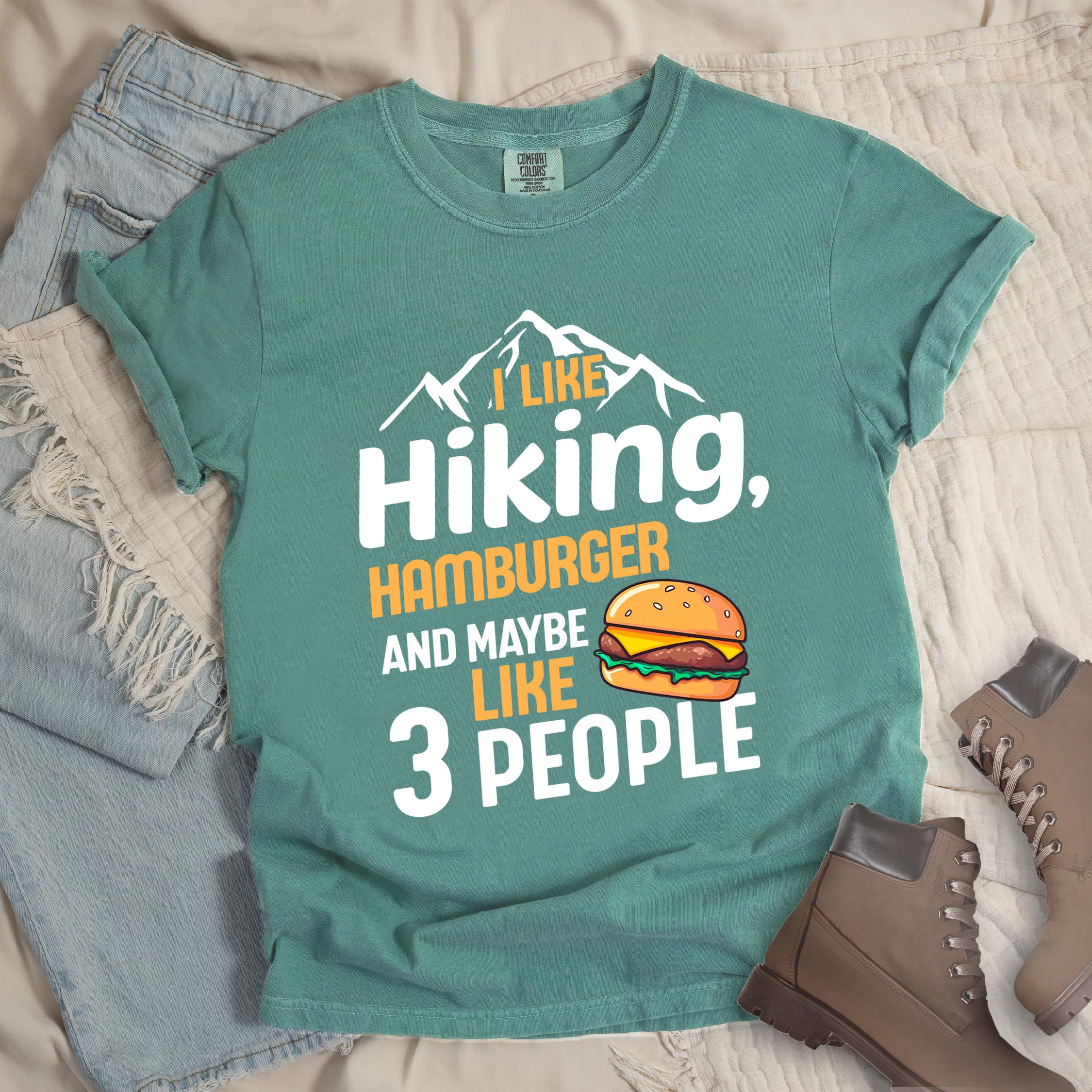 Green t-shirt with text "I LIKE Hiking, HAMBURGER AND MAYBE LIKE 3 PEOPLE". mountain silhouettes appear at the top. An illustration of a hamburger with sesame seed bun, cheese, lettuce and patty appears next to the word "LIKE". The design combines hiking and introvert humor.