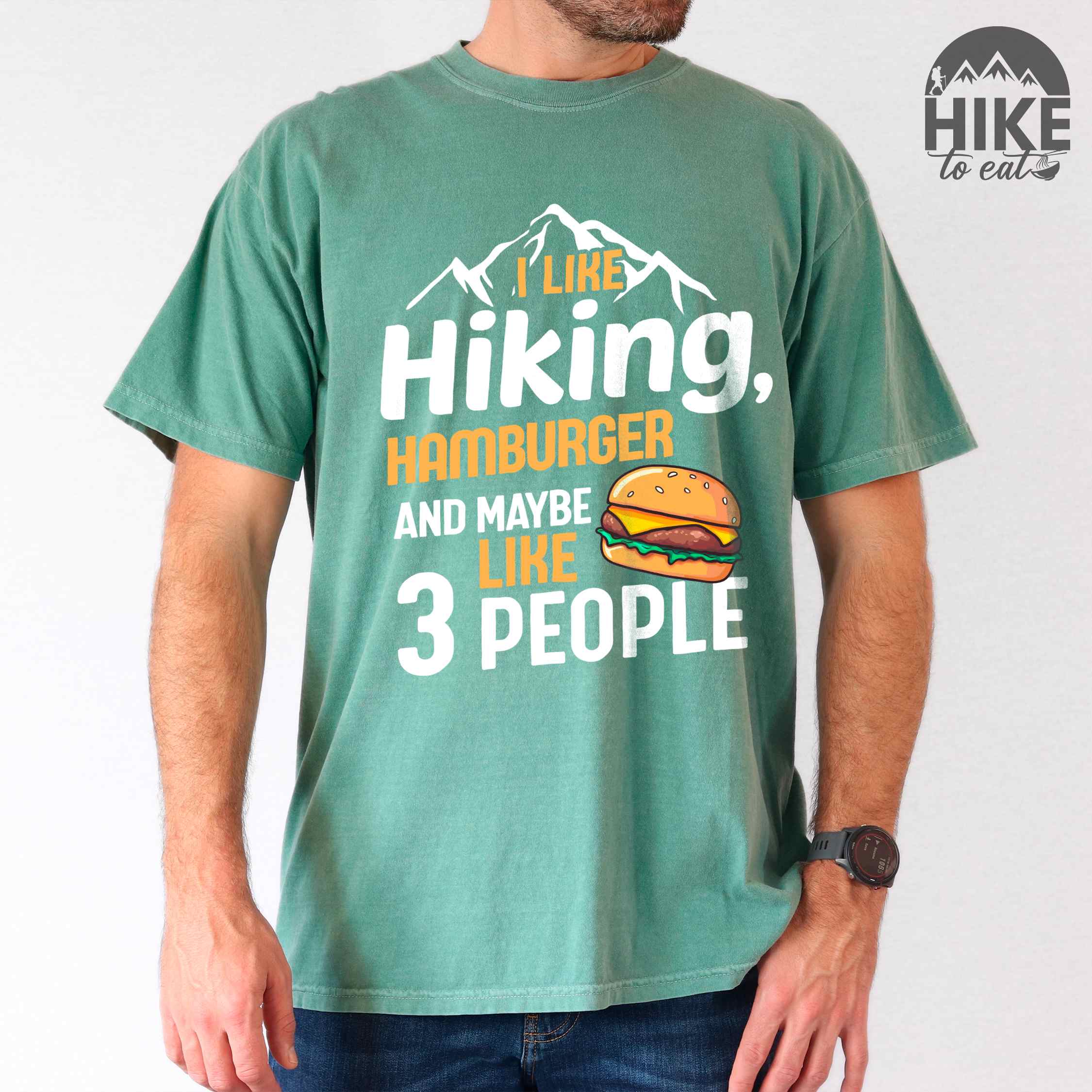 Man wearing light green "I like hiking, hamburger and maybe like three people" colored vintage tee with bold typography, birds, mountains, illustrated juicy hamburger, and backpacker for burger enthusiasts.